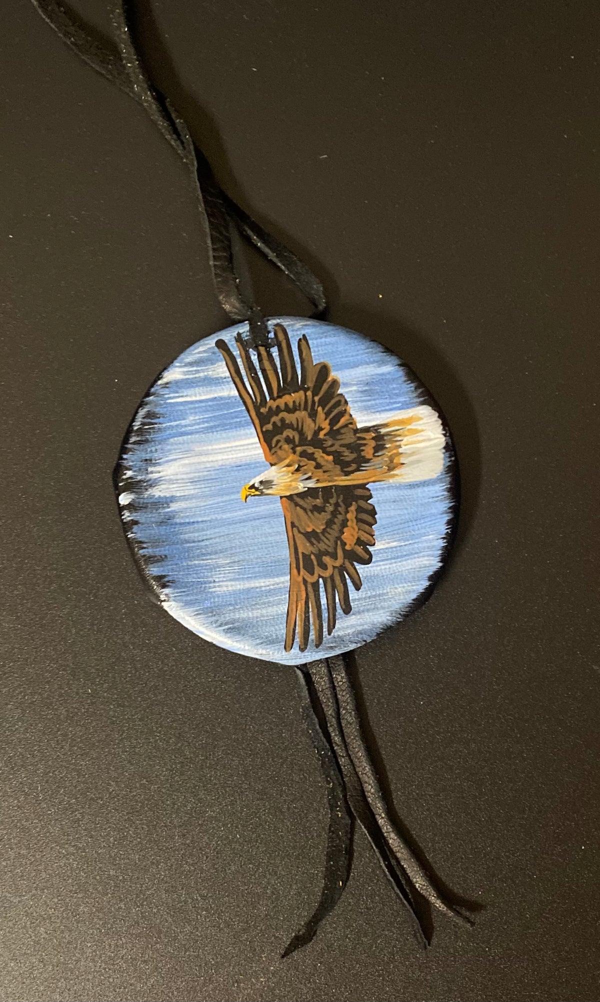 A circular leather shield featuring a hand-painted bald eagle in flight against a blue sky background, suspended from black leather ties on a dark brown surface.