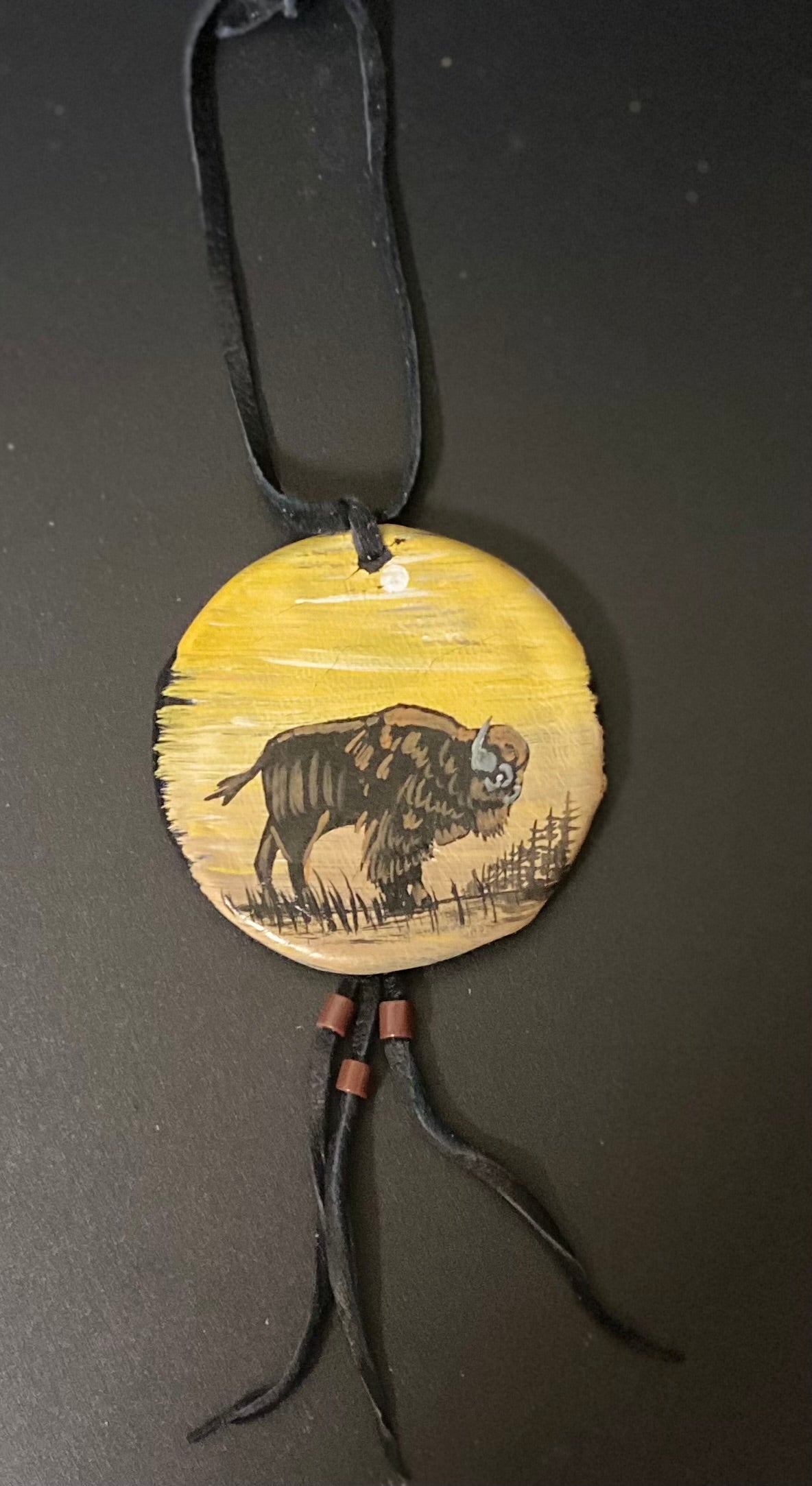 A circular shield featuring a hand-painted bison on a yellow background, suspended from a black cord with small brown beads, set against a dark gray background.