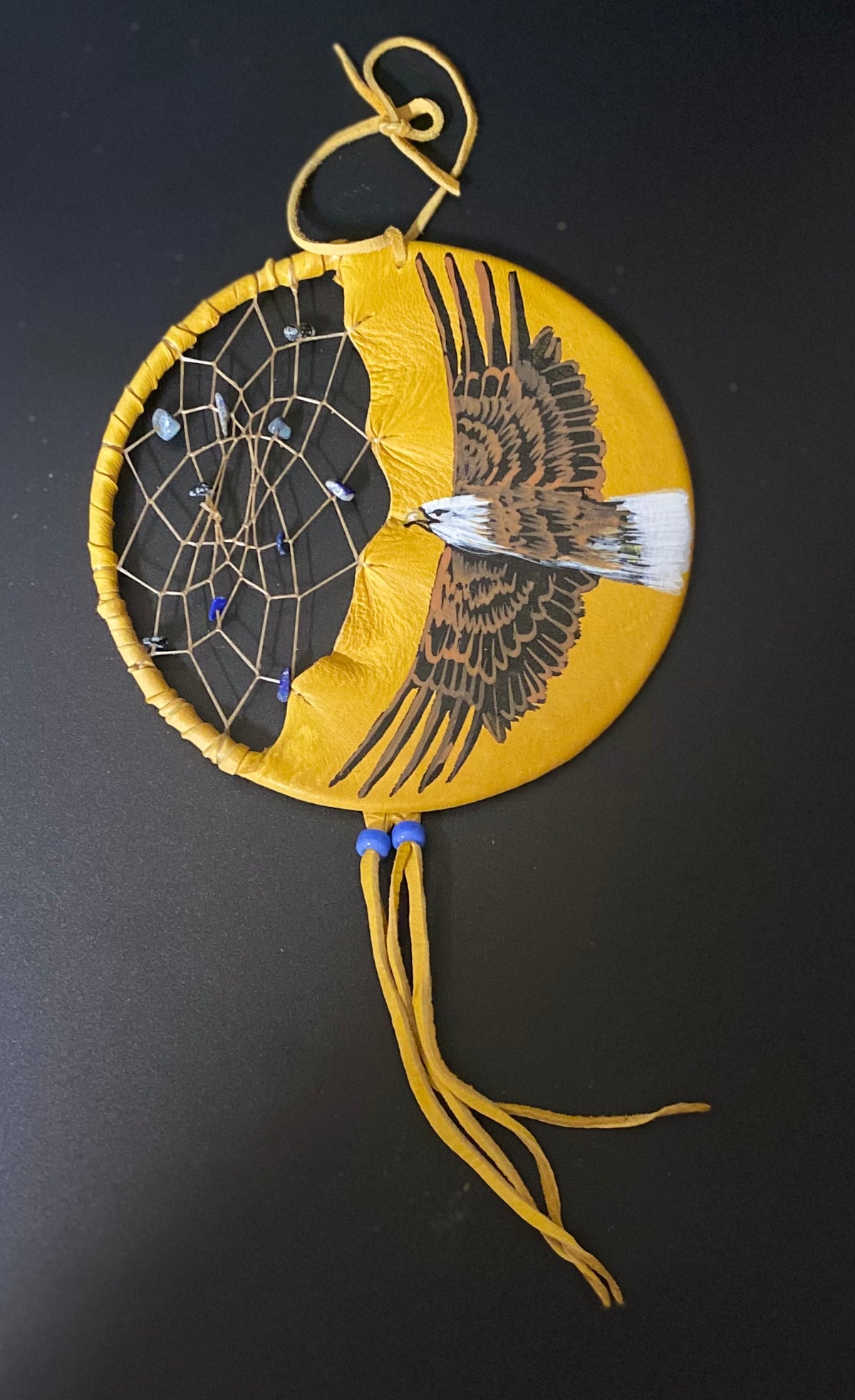 A circular handmade dreamcatcher with a yellow leather body, featuring a painted bald eagle on one side and a web pattern on the other, hanging against a dark background.