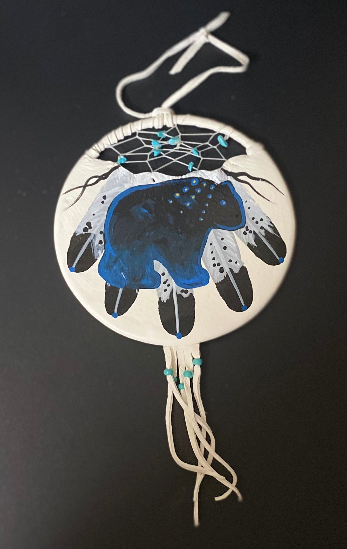 A circular Native American style dreamcatcher wall hanging with a hand-painted bear design in blue, black, and white, featuring a white woven web and hanging cords against a dark background.