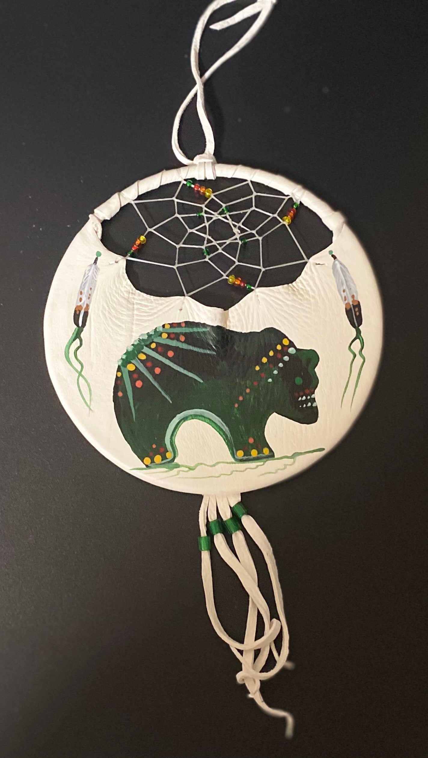 A circular handmade dreamcatcher ornament featuring a painted green bear and spider design on a dark background, with white hanging cords and small bead accents, photographed against a dark backdrop.