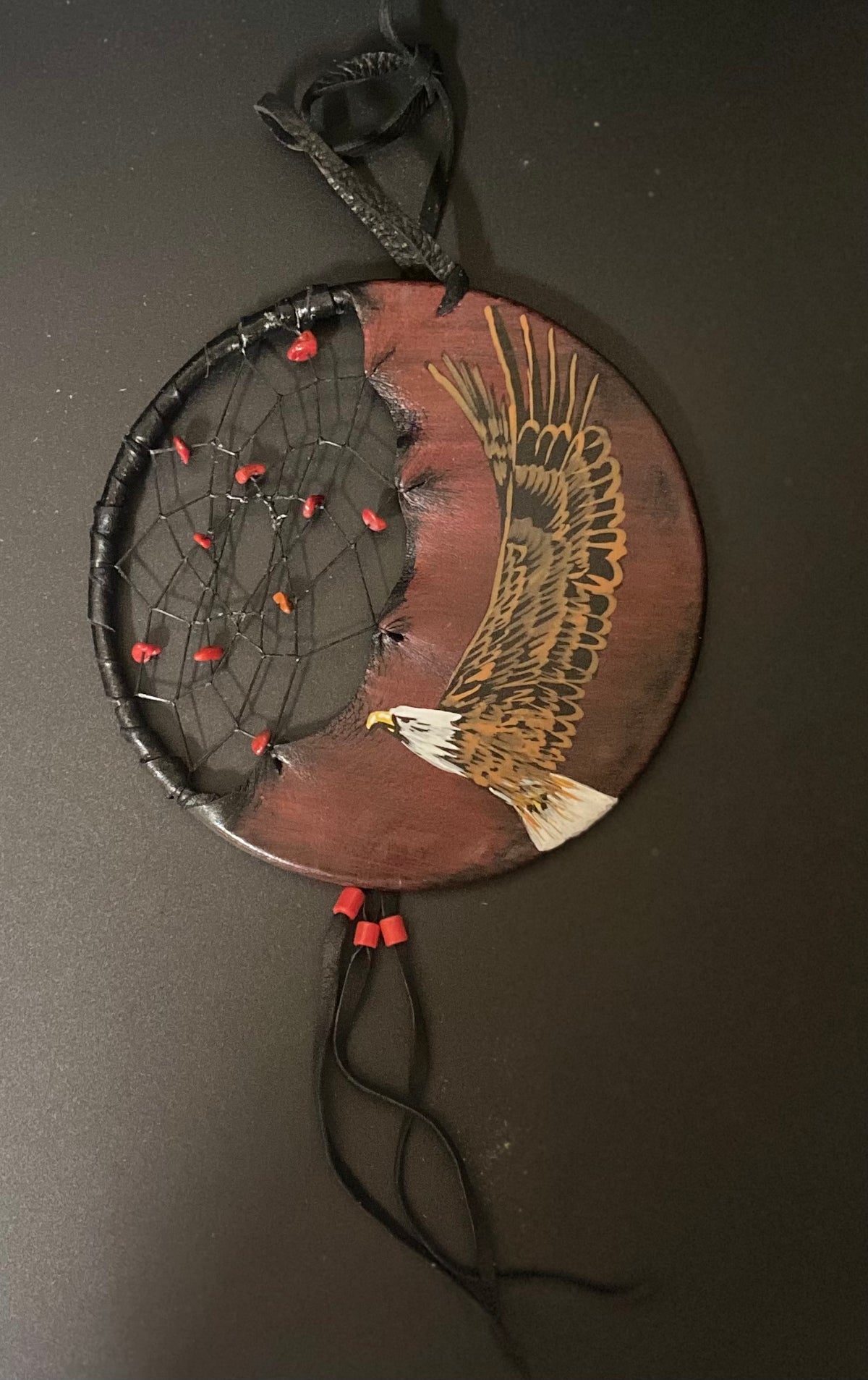 A circular dreamcatcher wall hanging with a painted bald eagle on one side and a black webbed center with small red beads, shown against a dark background.