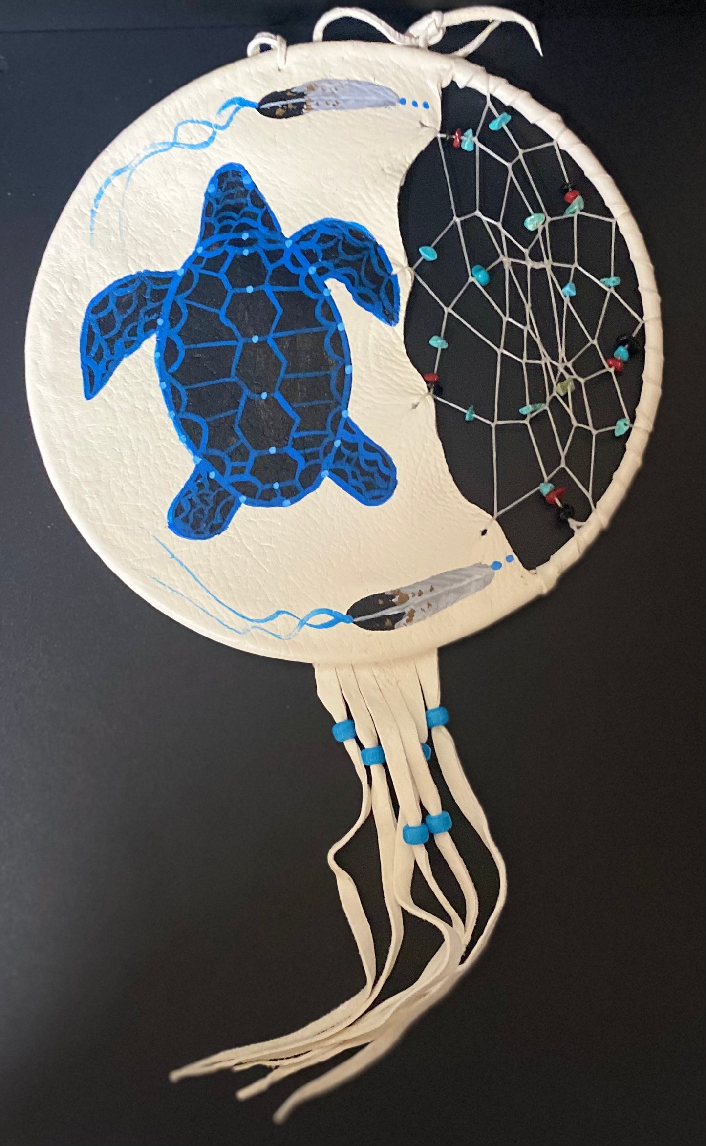 A circular handmade dreamcatcher wall hanging with a blue sea turtle painted on the left side, a black webbed center with small turquoise and red beads, cream-colored hanging cords, and a white hanging loop at the top, shown against a dark background.