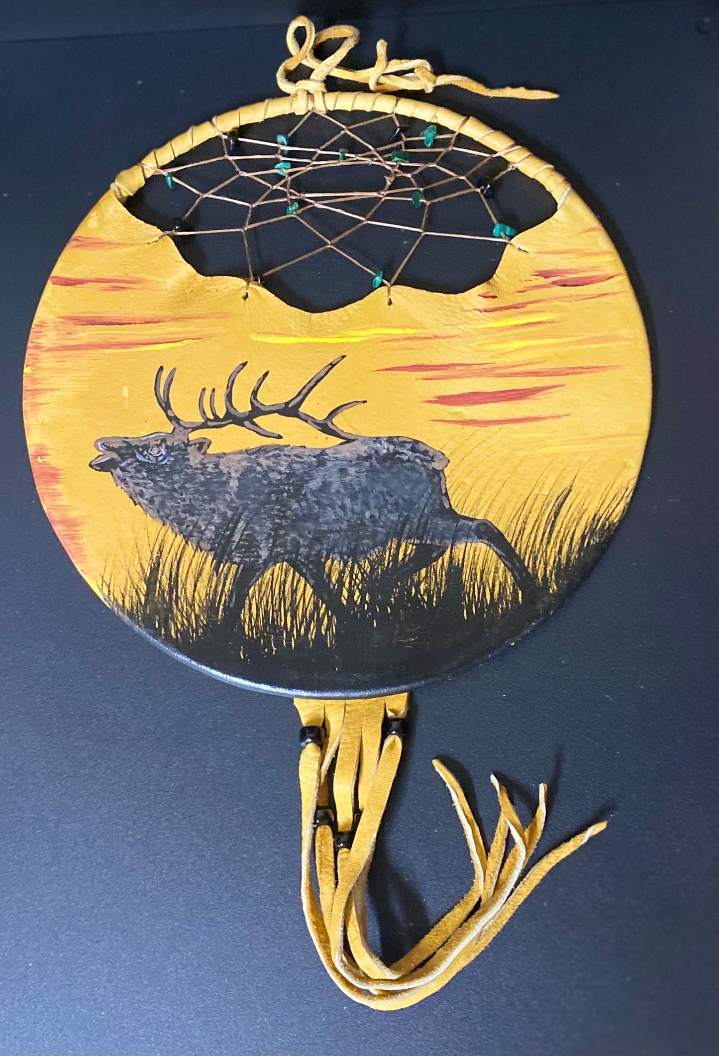 A circular handcrafted dreamcatcher wall hanging with a painted elk silhouette and sunset design, featuring tan leather cords and a hanging loop at the top, photographed against a dark background.