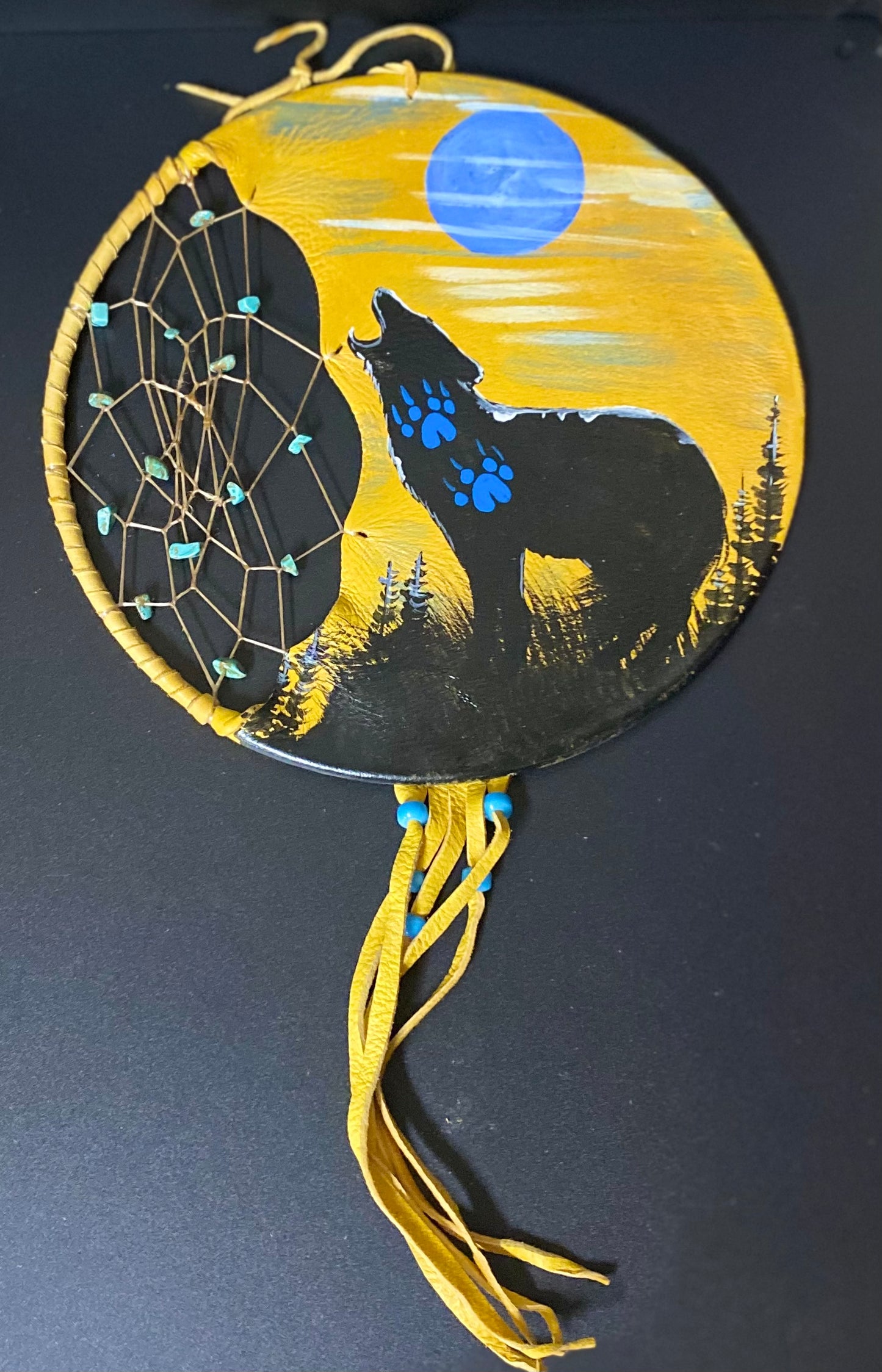 A circular dreamcatcher wall hanging with a painted wolf howling at a full moon. The design features a black wolf silhouette, a blue moon, and a yellow background, with a woven web on the left and yellow hanging tassels below.