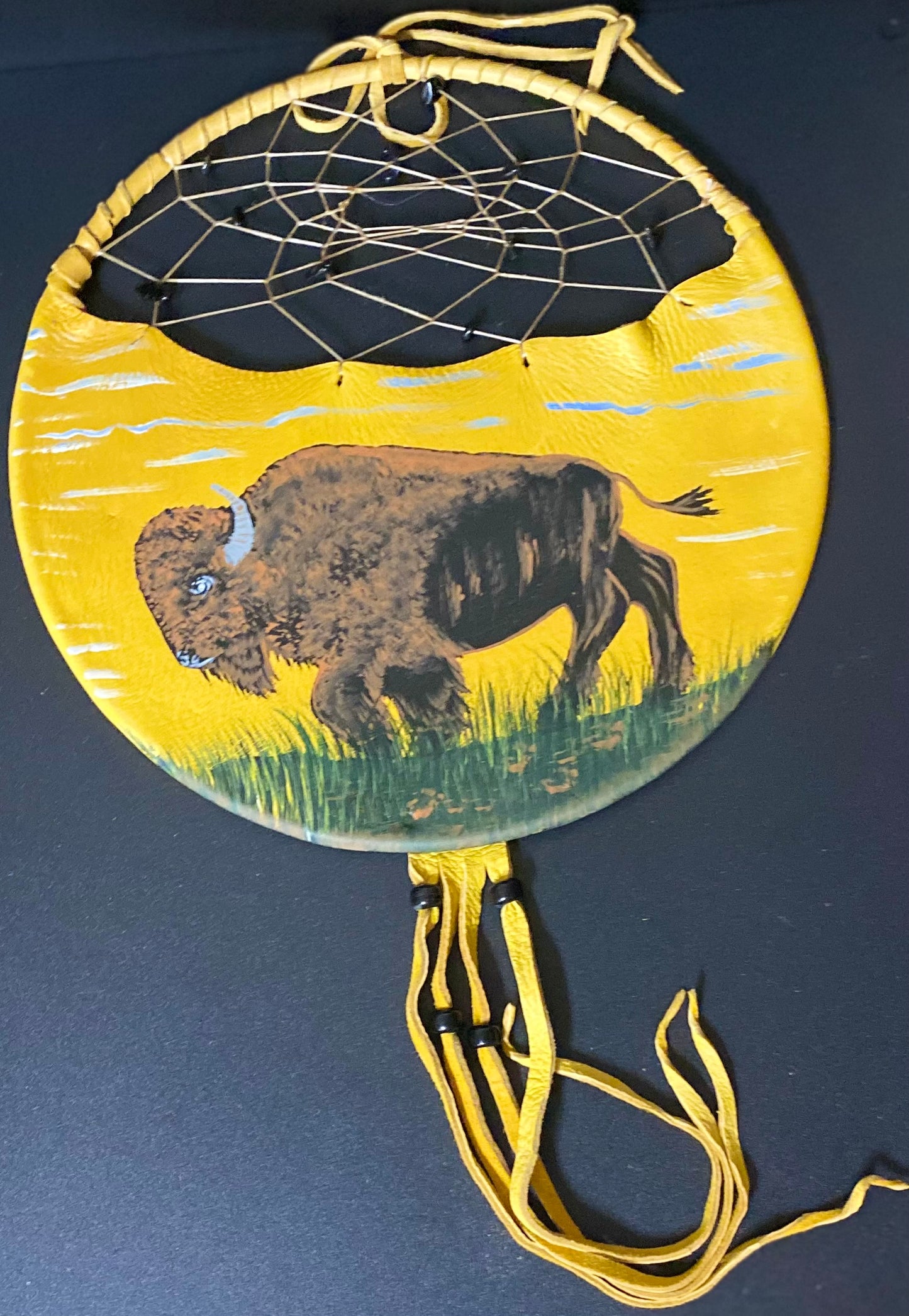 A circular hand-painted Native American style dreamcatcher with a yellow leather body, painted bison design, and yellow webbing, shown on a dark background.