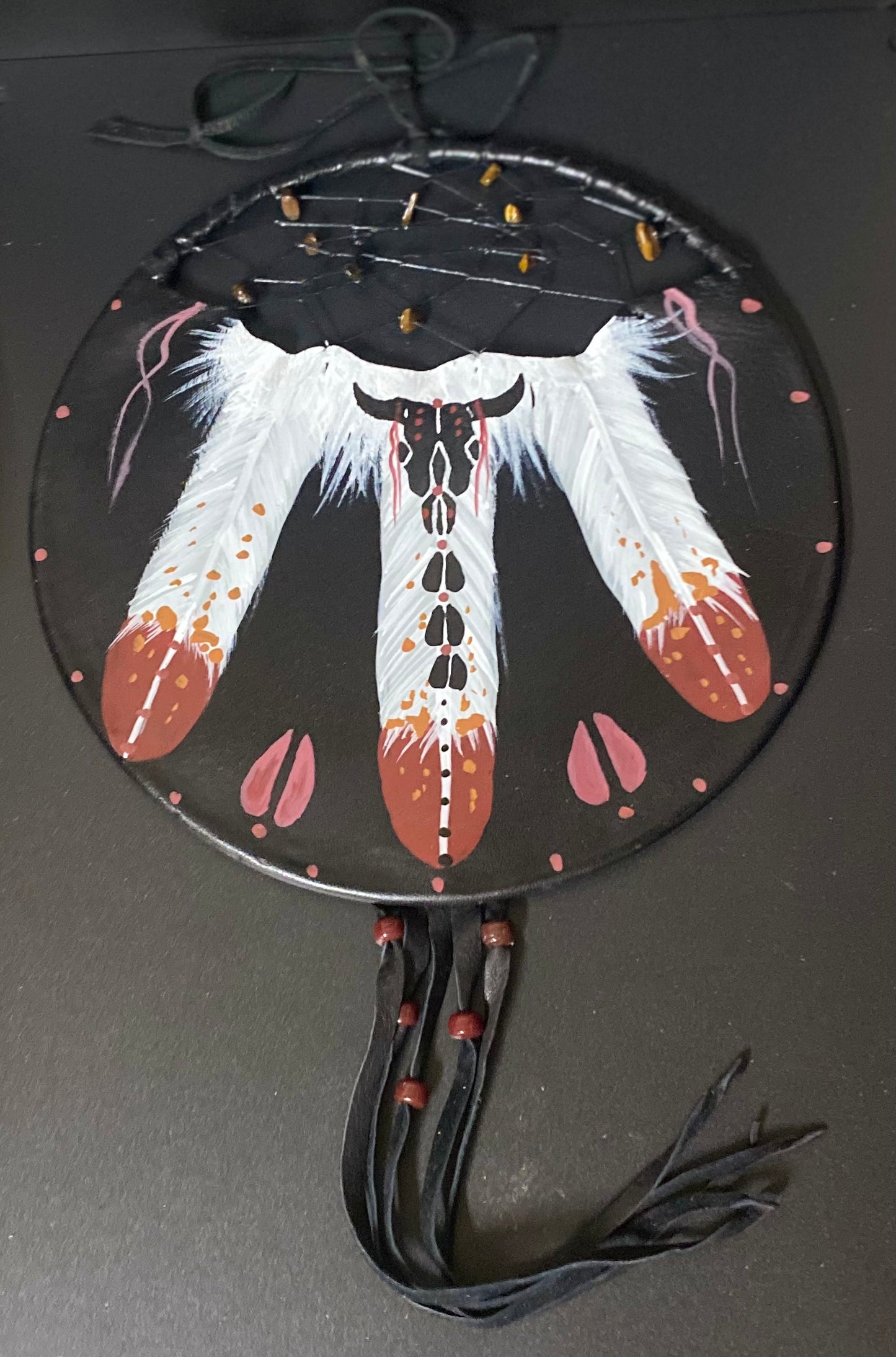 A circular black dreamcatcher-style wall hanging or decorative piece with a large white feather design, pink and orange painted accents, black hanging cords, and small bead details on a dark background.