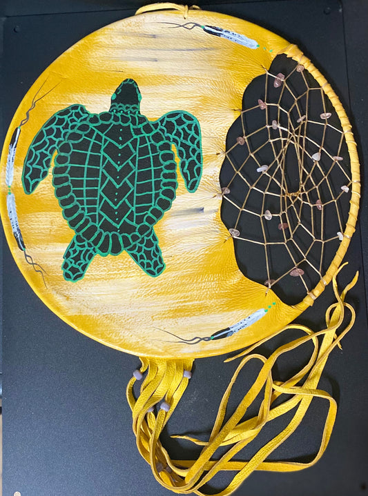 A circular handmade dreamcatcher wall hanging with a yellow painted background, a green sea turtle design on the left, a black webbed center section on the right, and multiple yellow hanging cords below, photographed against a dark surface.