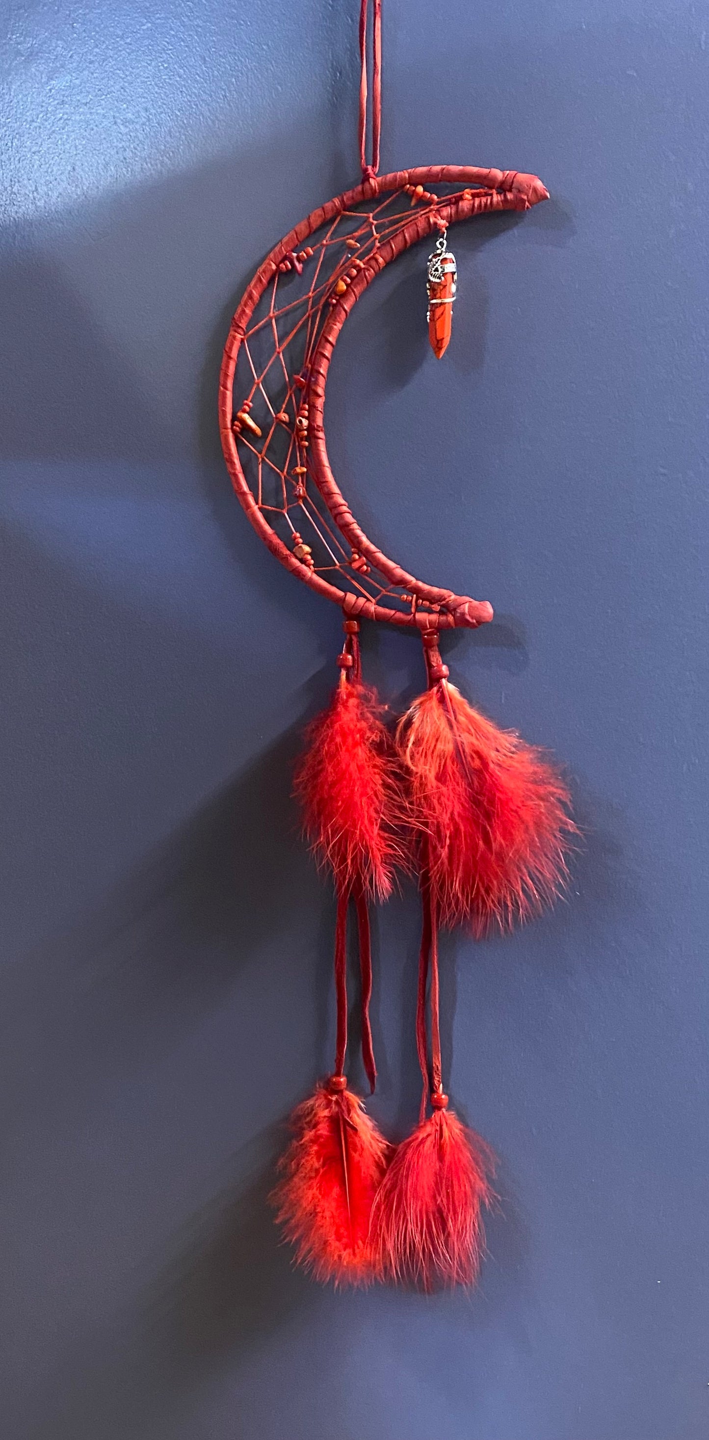 A red crescent moon dreamcatcher hanging against a dark blue wall, featuring wrapped red cord, small bead details, and three hanging red feathers.