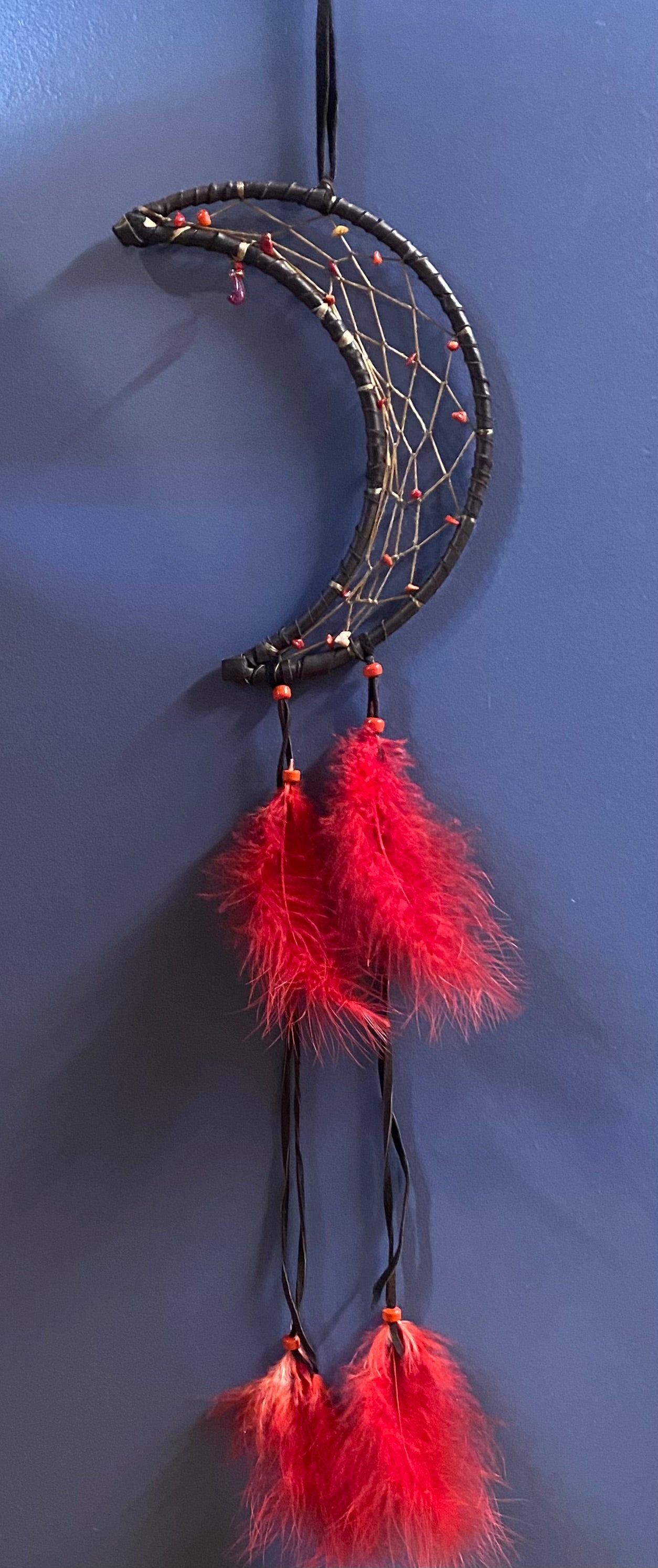 A crescent moon dreamcatcher with a black wrapped frame, white webbing, and bright red feathers hanging against a dark blue wall.