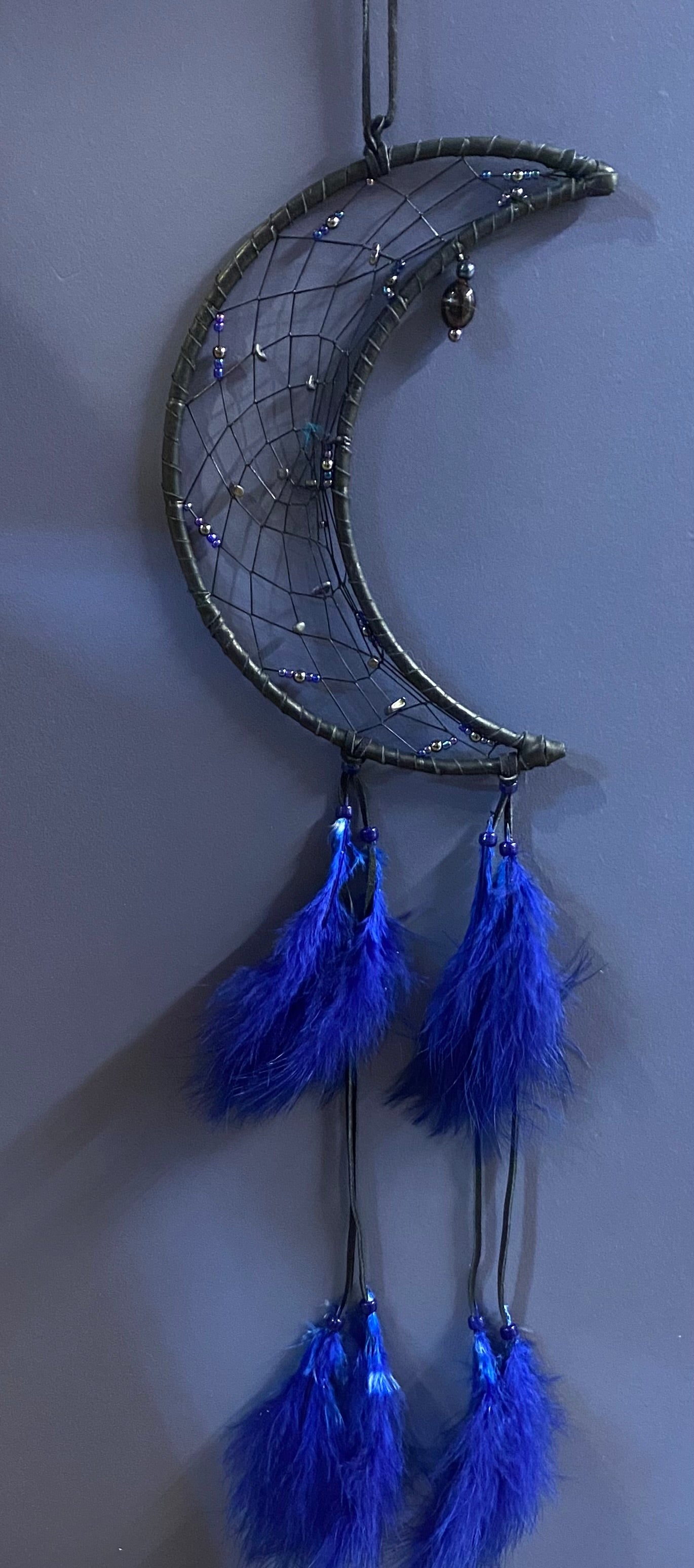 A crescent moon dreamcatcher with a dark metal frame, wrapped in dark gray webbing with small reflective beads, and hanging four bright royal blue feathers on black cords against a plain gray wall.