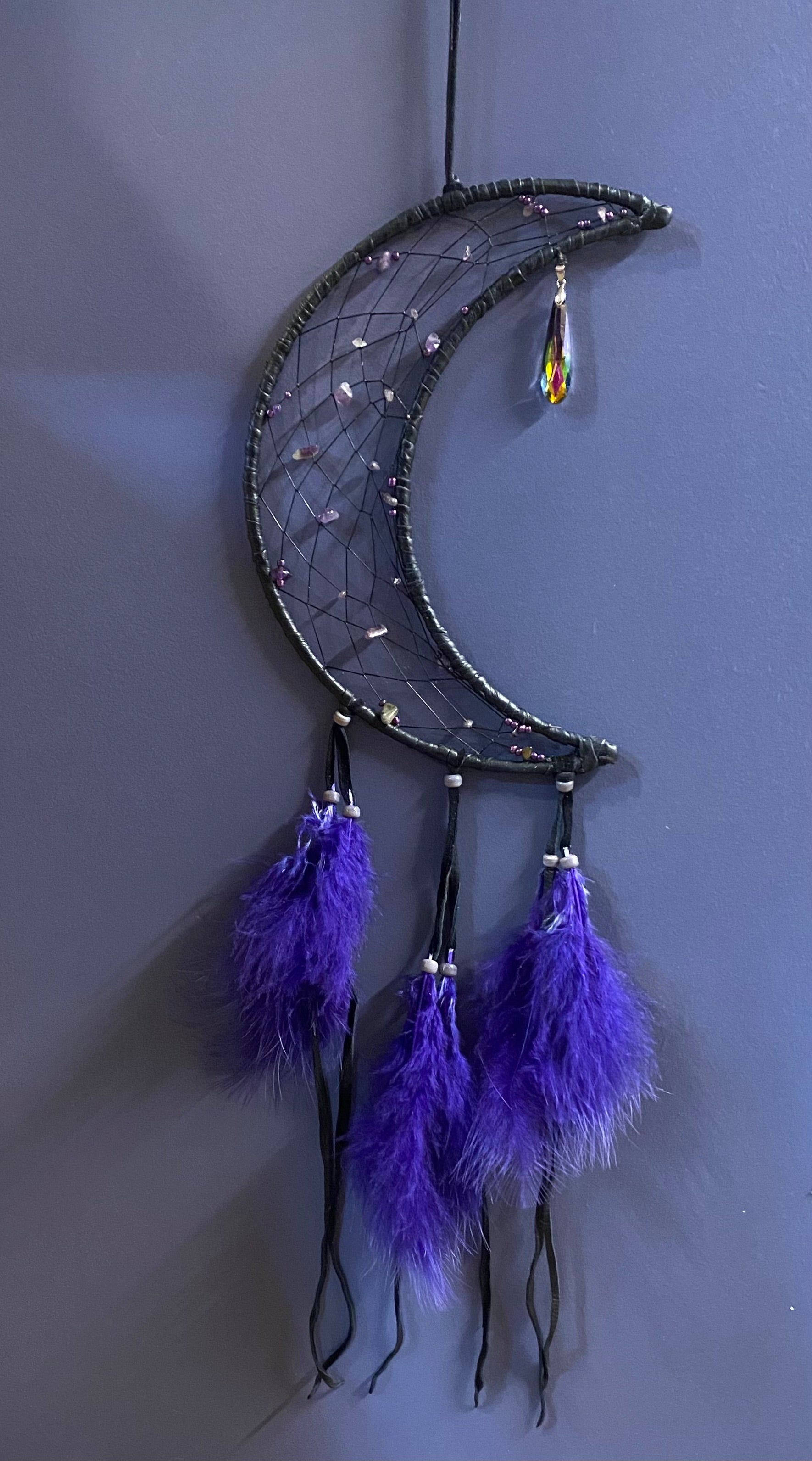 A crescent moon dreamcatcher hanging against a dark gray wall. The moon frame is dark gray with a black web, decorated with small silver and purple beads, and has three purple feathers hanging from the bottom with black tassels.