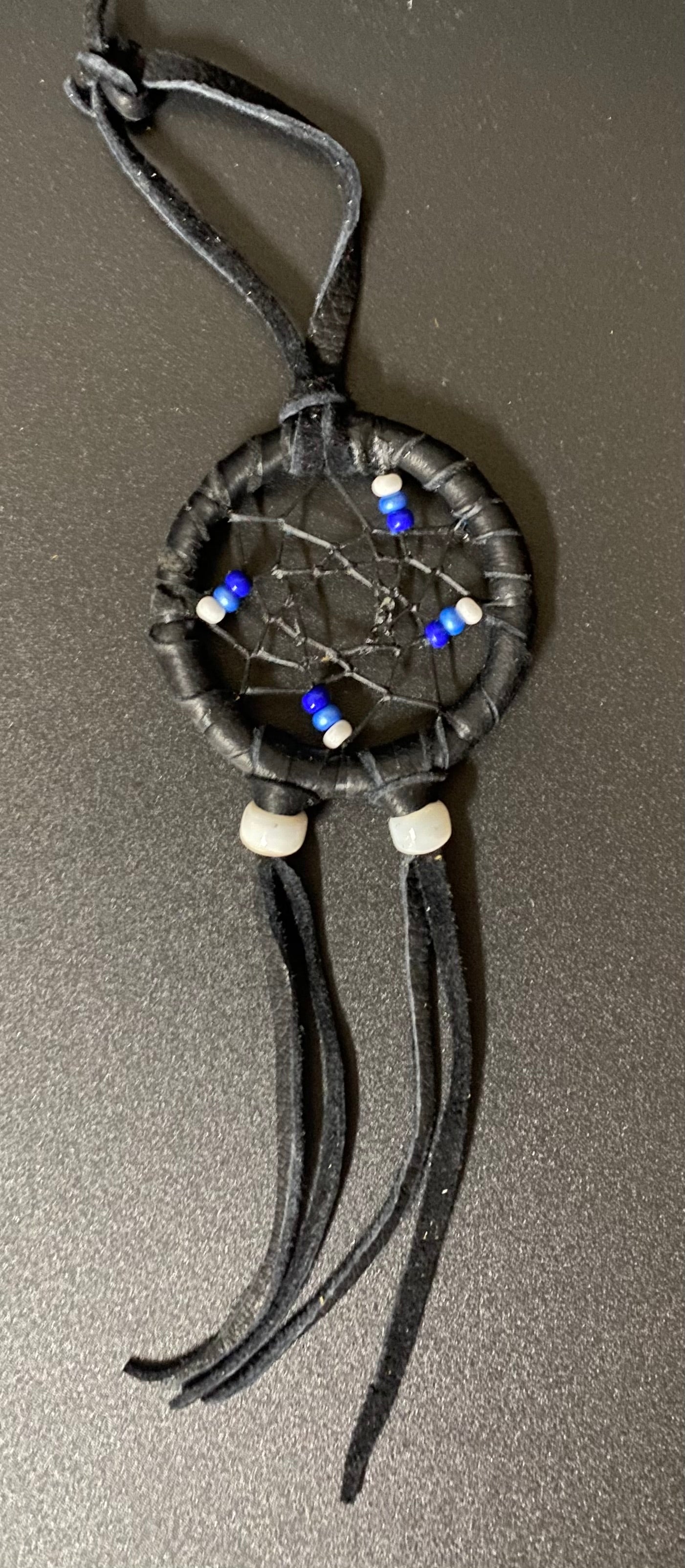 A small black leather dreamcatcher with a webbed center, decorated with blue and white beads, resting on a dark gray textured surface.