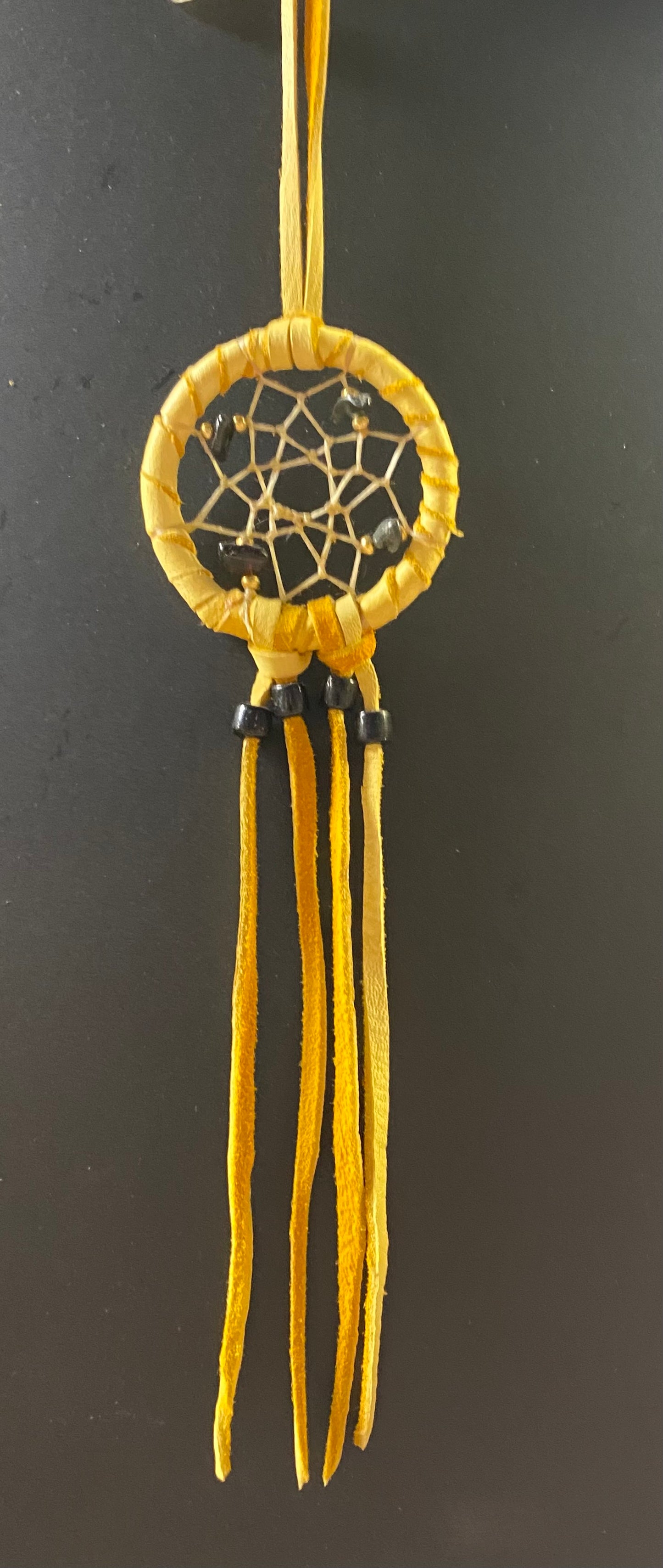A small yellow dreamcatcher with a circular web design and long hanging tassels, shown against a dark gray background.