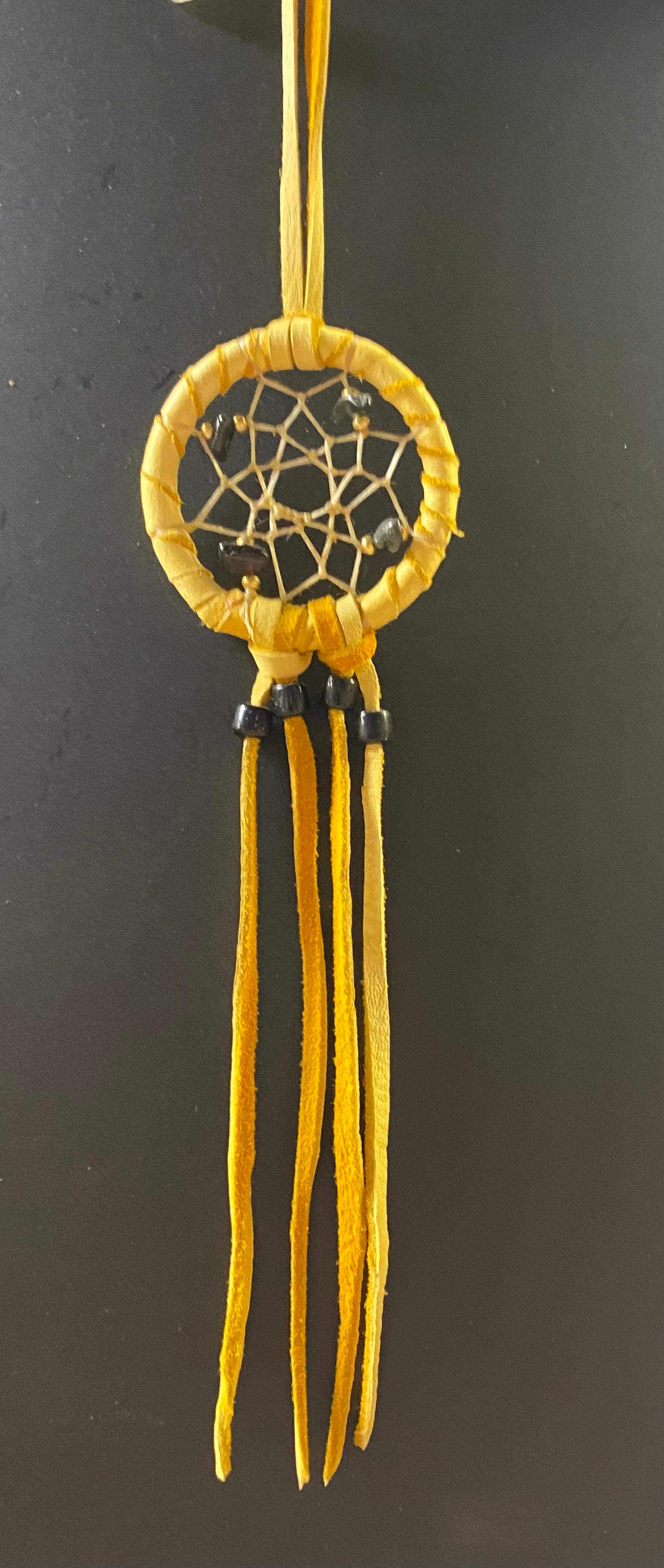 A small yellow dreamcatcher with a circular web design and long hanging tassels, shown against a dark gray background.