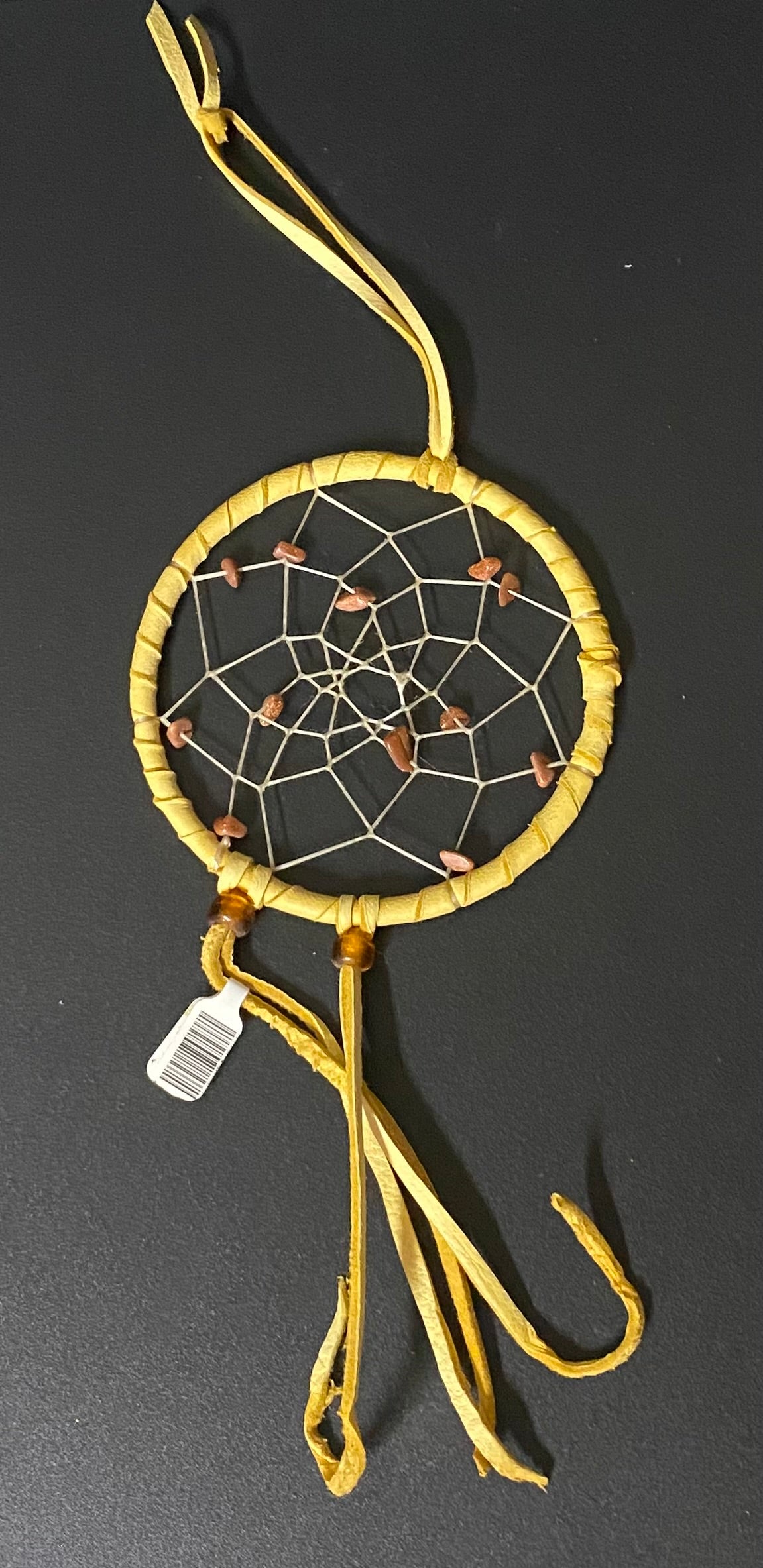 A small handmade dreamcatcher with a yellow wrapped hoop, white webbing, and brown beads, shown on a dark background with a small white tag attached.