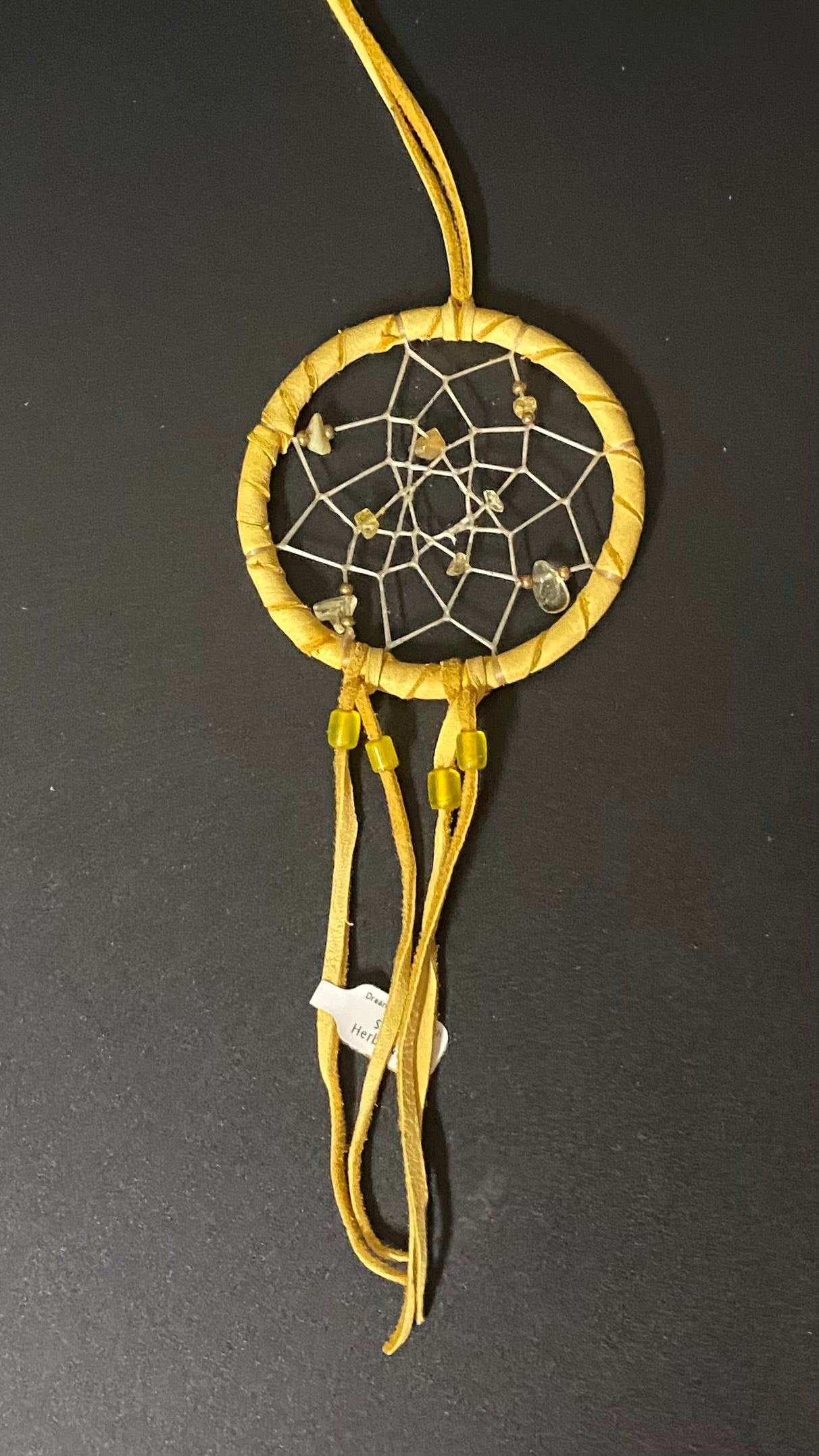 A small handmade dreamcatcher with a natural tan woven hoop, white webbing, and yellow cords hanging below, photographed against a dark gray background.