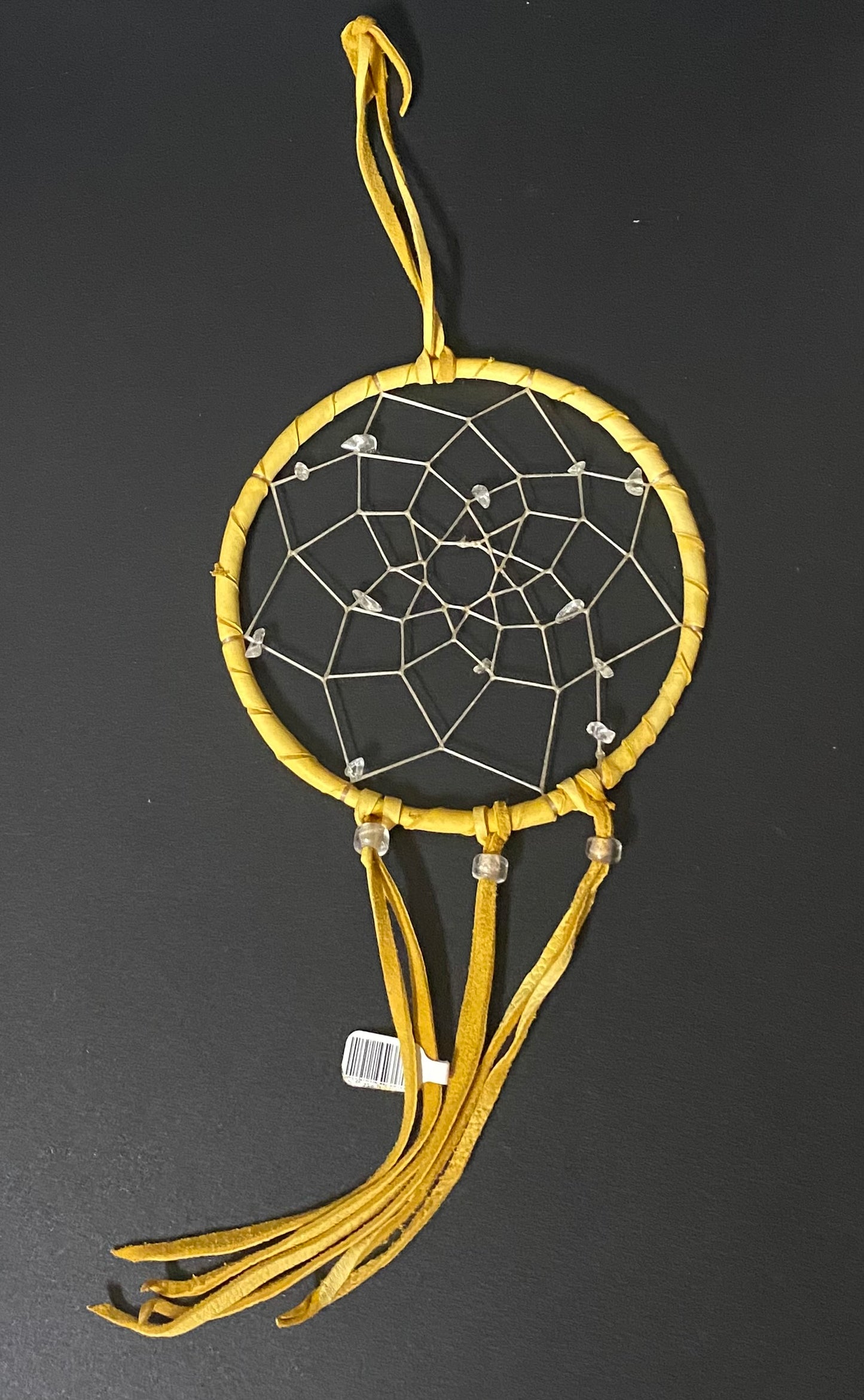 A yellow dreamcatcher with a wrapped circular frame, white webbing, and three hanging tassels, photographed against a dark gray background.
