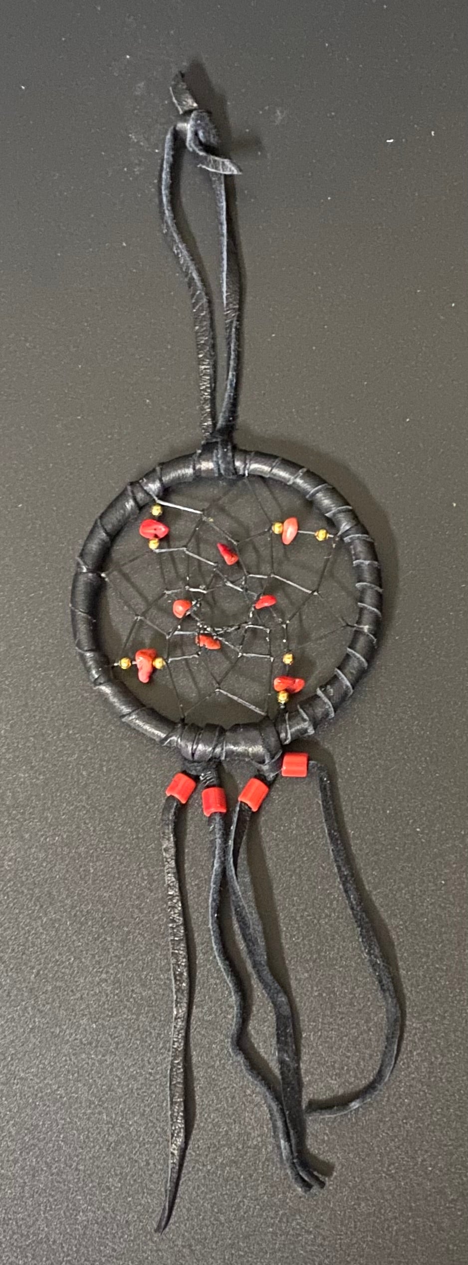 A handmade dreamcatcher with a dark gray wrapped hoop, black webbing, and hanging black leather strands, decorated with small red and orange beads.