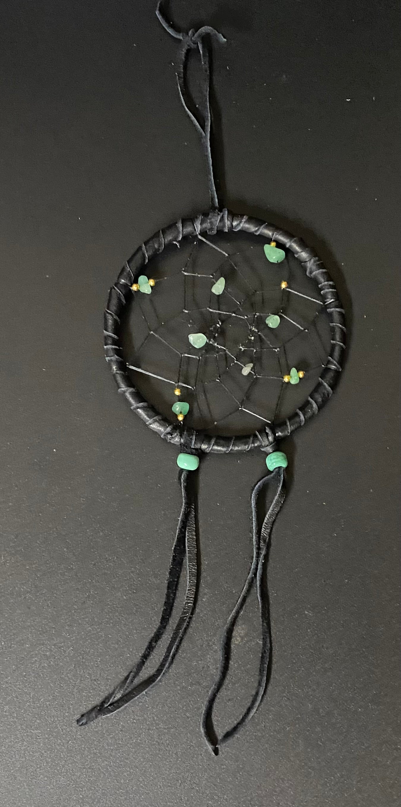 A black dreamcatcher with wrapped black cords, a webbed center, small green and yellow beads, and three hanging black tassels, photographed against a dark gray background.