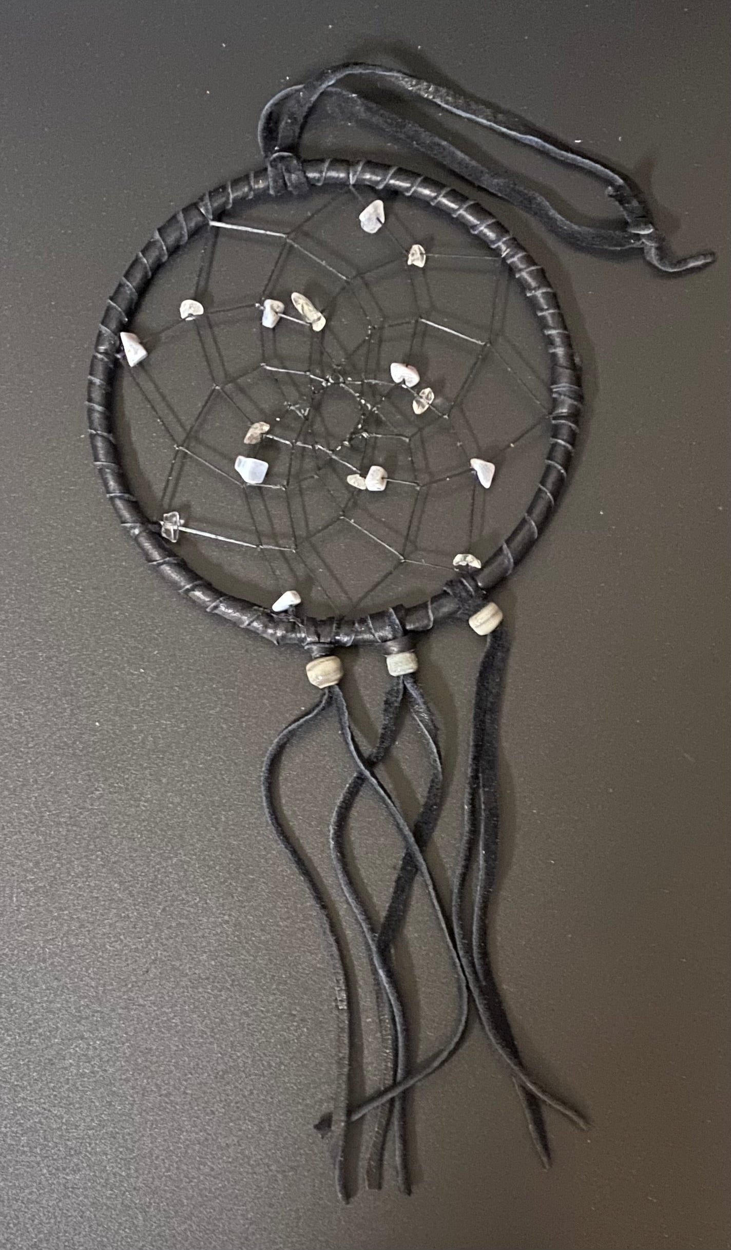 A black leather dreamcatcher with small white stones and beads, resting on a dark gray surface.