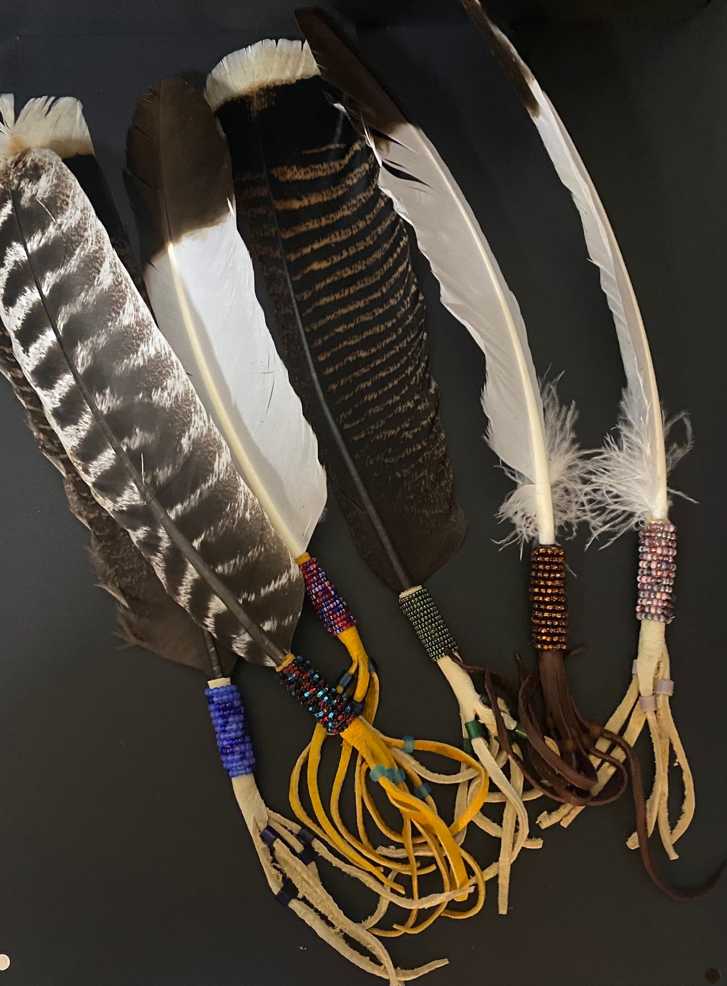 Four decorative feathers laid flat on a dark background, each with natural brown and white plumage and colorful beaded bands near the base, with tan cords wrapped around the ends.