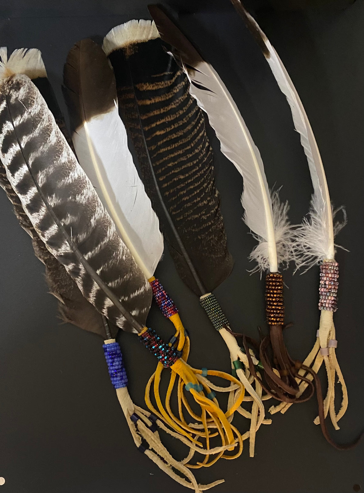 Four decorative feathers laid flat on a dark background, each with natural brown and white plumage and colorful beaded bands near the base, with tan cords wrapped around the ends.