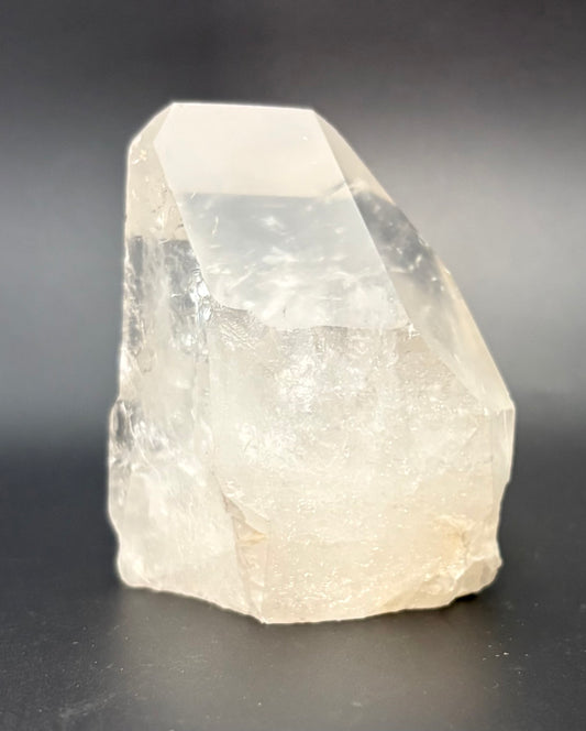 A polished quartz crystal with a pointed geometric shape, made of semi-translucent off-white material, photographed on a dark gray background.