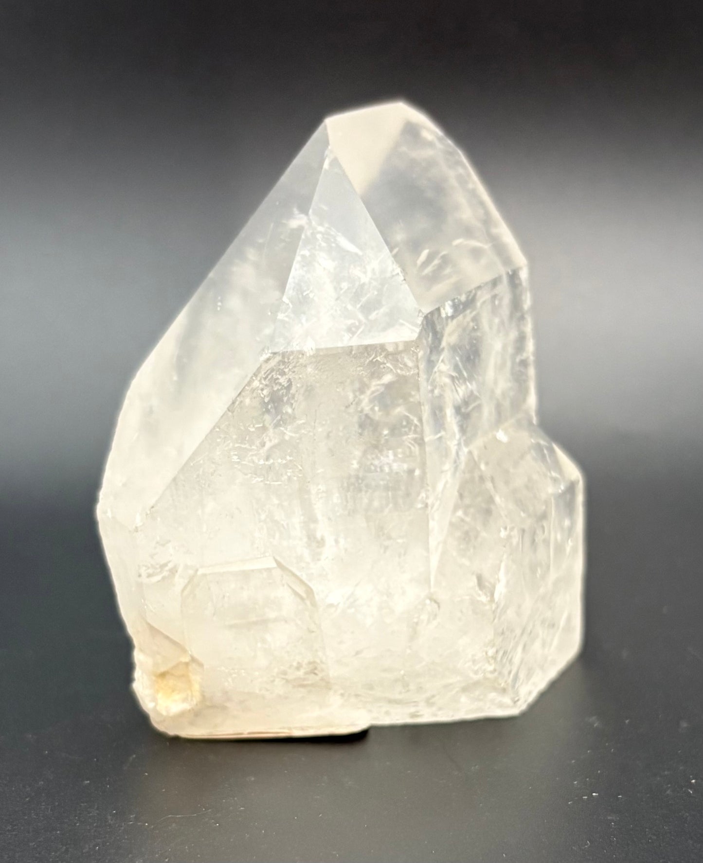 A polished quartz crystal with a pointed geometric shape, made of semi-translucent off-white material, photographed on a dark gray background.
