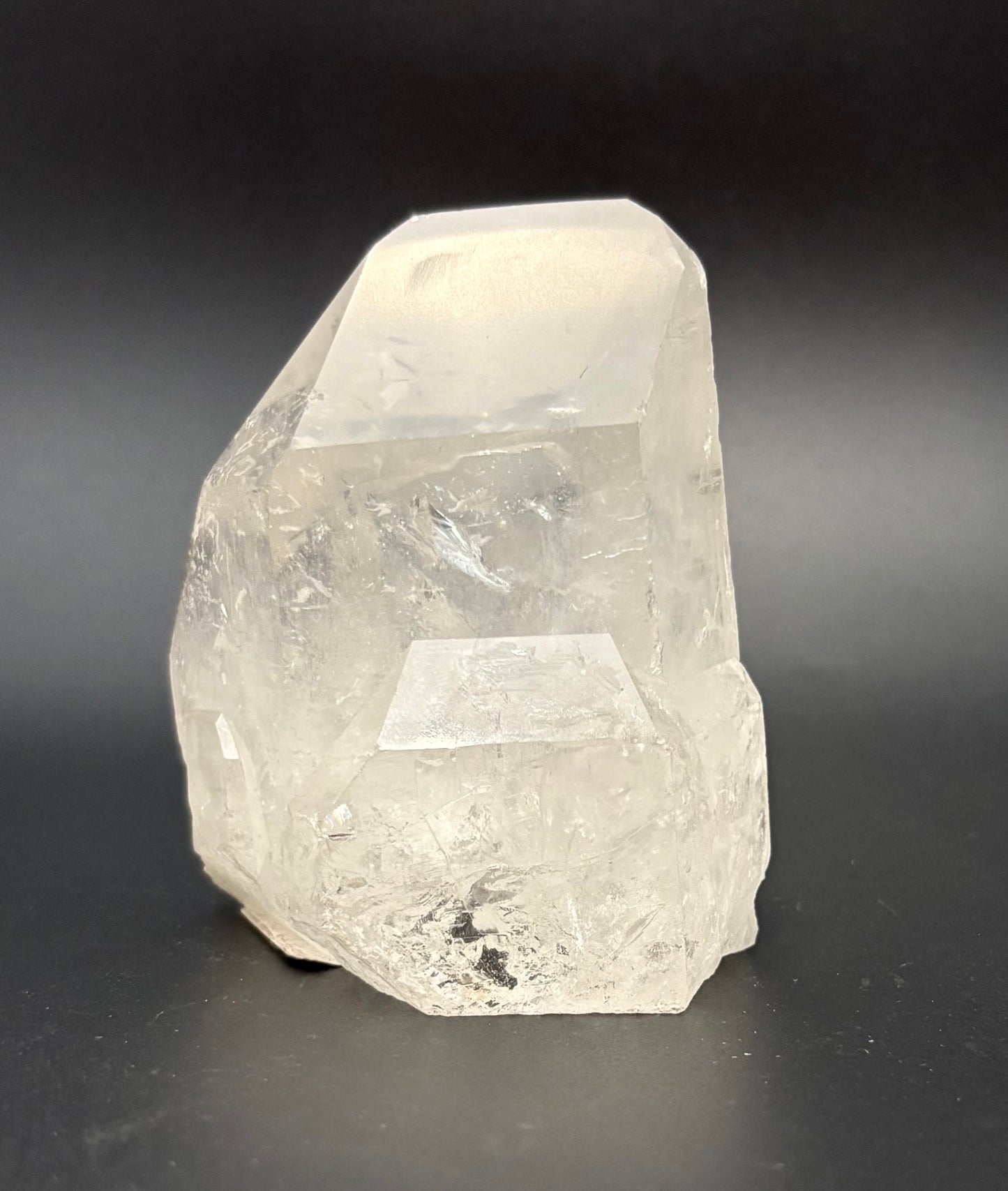 A raw, unpolished clear quartz crystal with natural faceted edges, standing upright on a dark gray to black background.