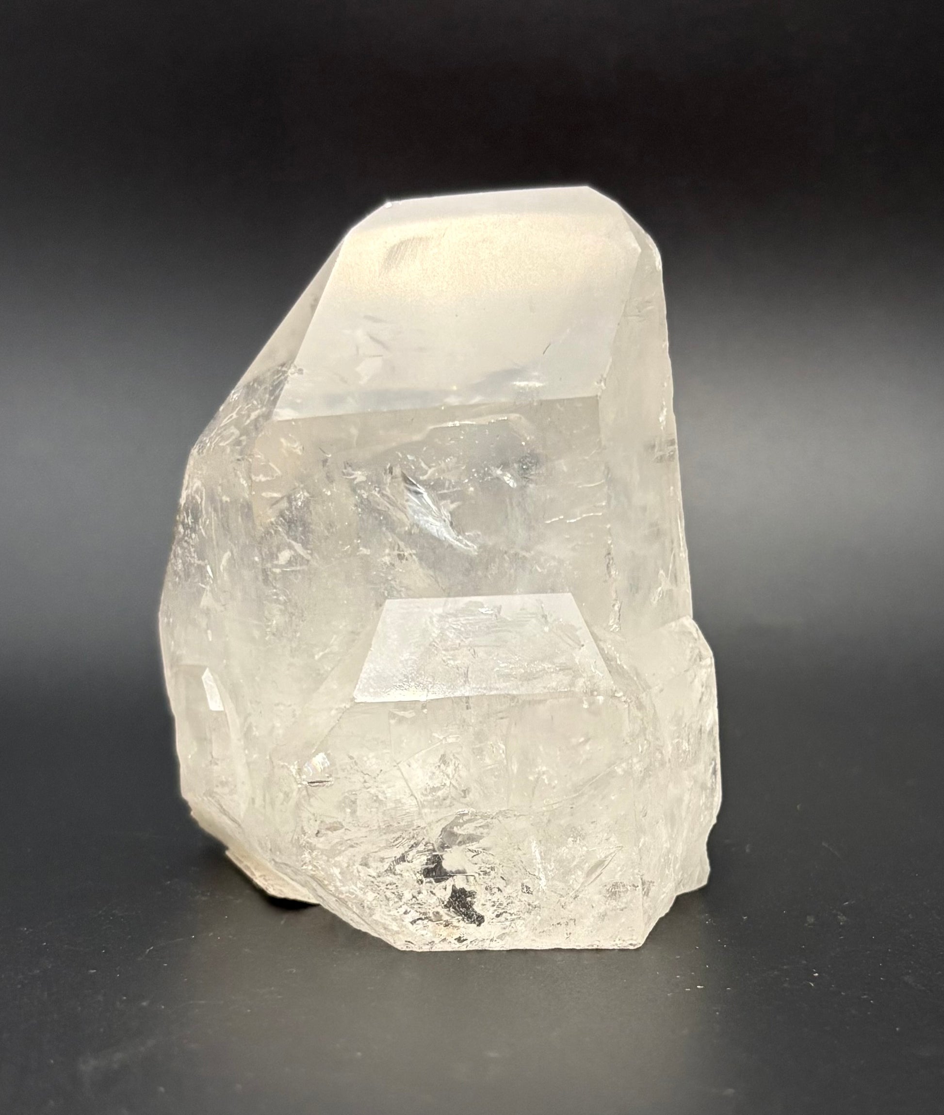 A raw, unpolished clear quartz crystal with natural faceted edges, standing upright on a dark gray to black background.