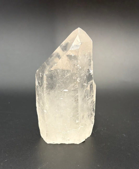 A raw, natural clear quartz crystal point with visible internal inclusions, standing on a dark surface against a dark gray gradient background.