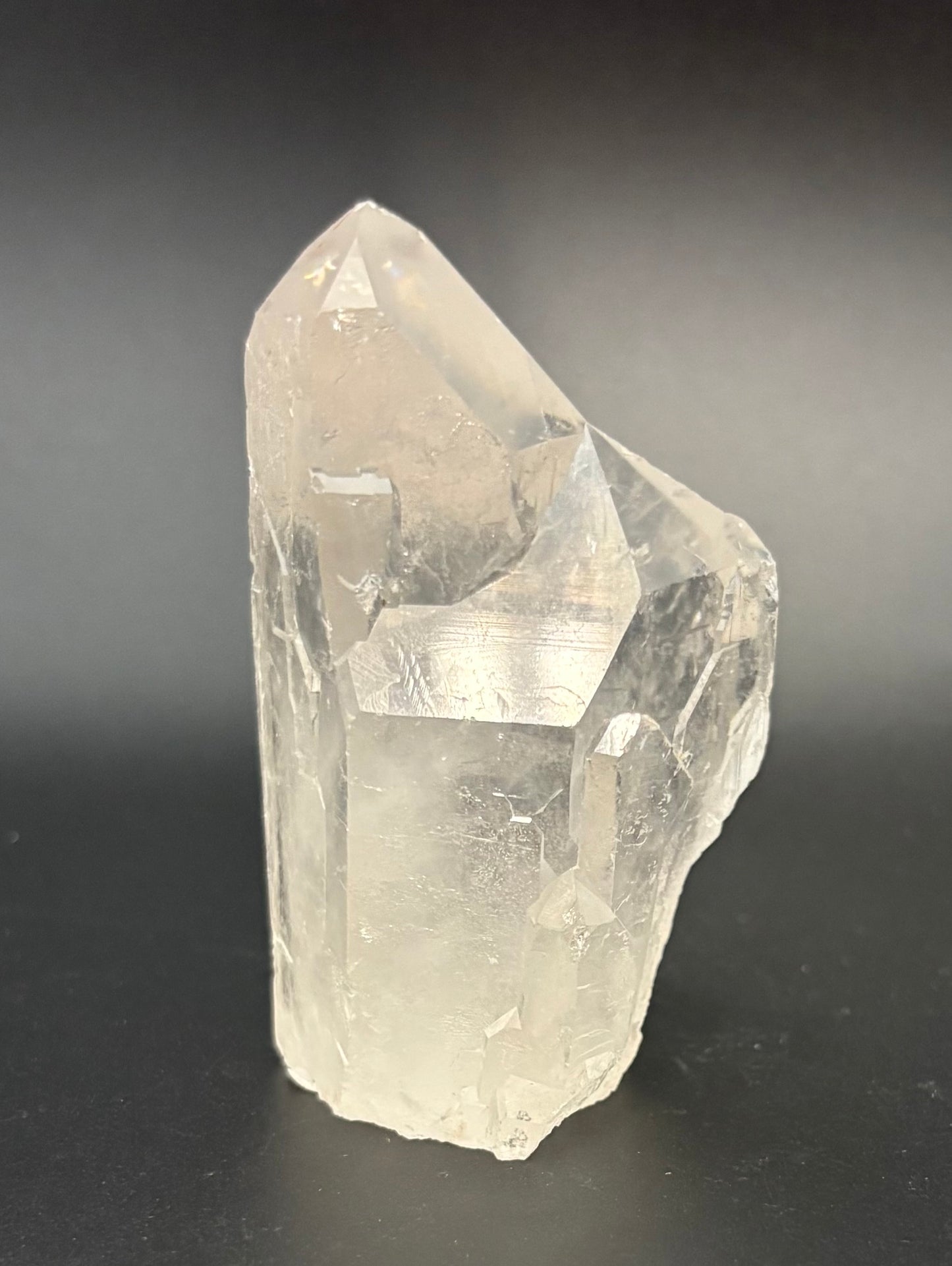 A raw, unpolished clear quartz crystal cluster with natural geometric facets, standing upright on a dark gray to black background.