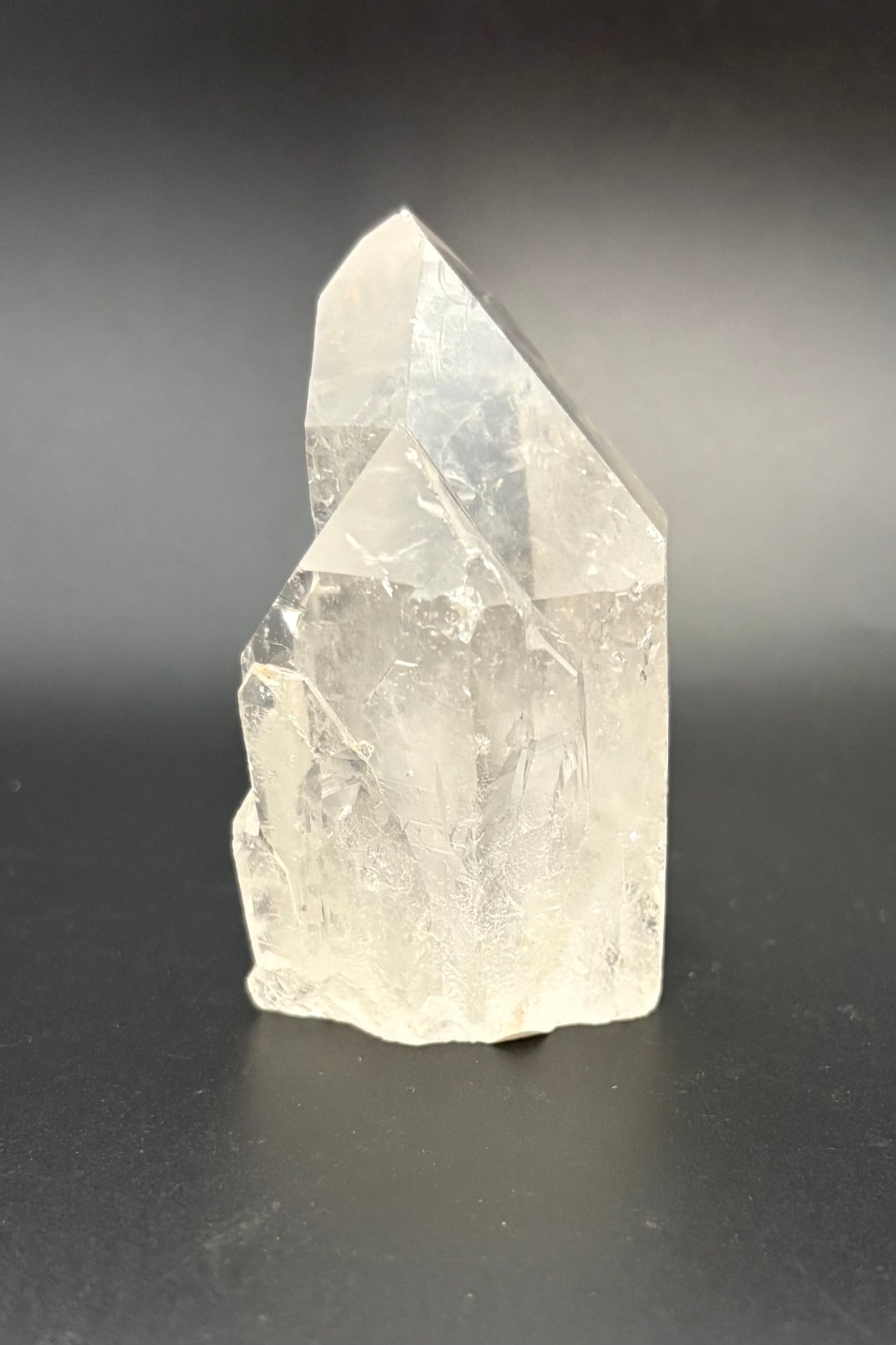 A raw, unpolished clear quartz crystal cluster with natural geometric facets, standing upright on a dark gray to black background.
