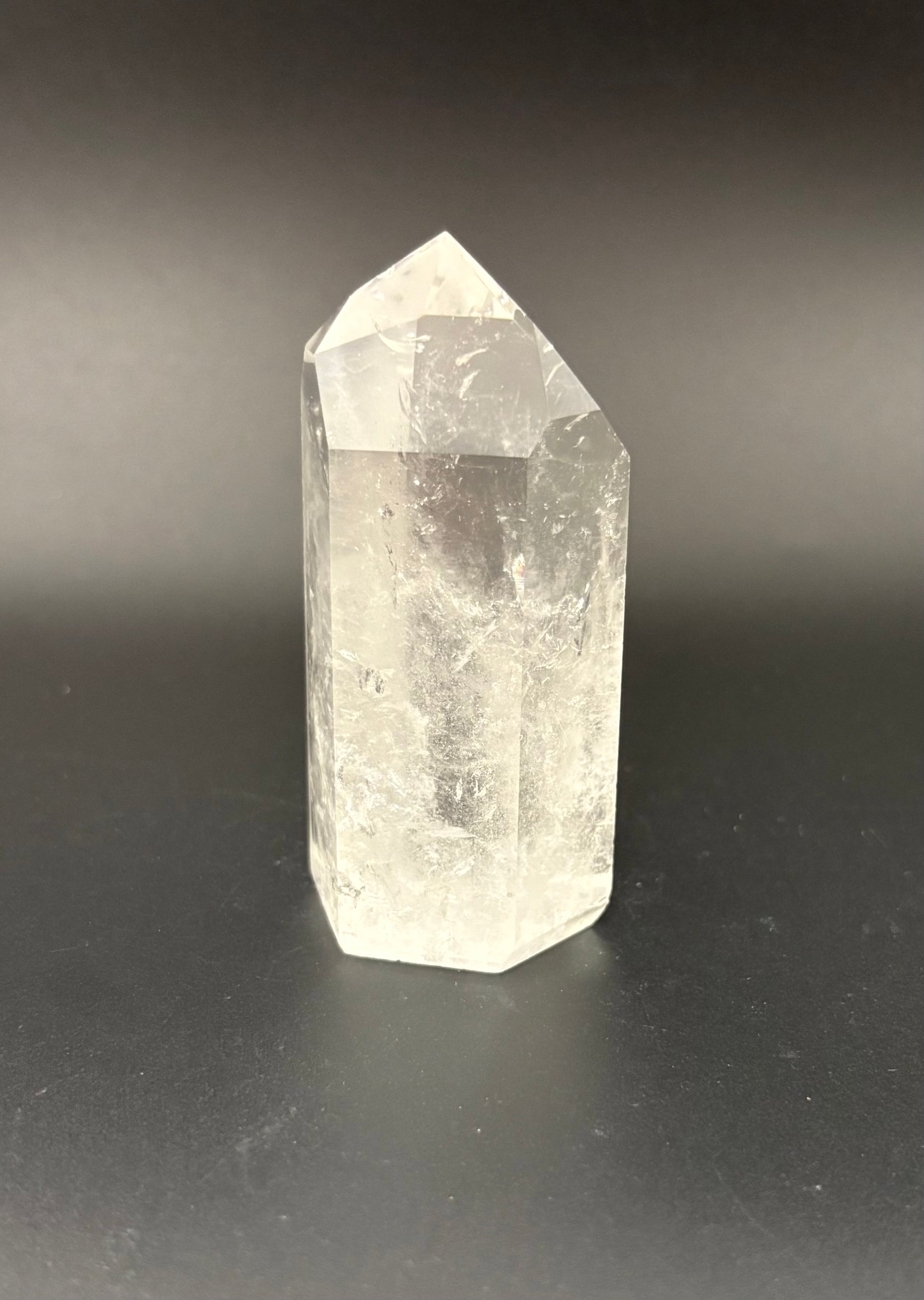 A natural clear quartz crystal point tower standing upright on a dark gray surface against a dark gray background.