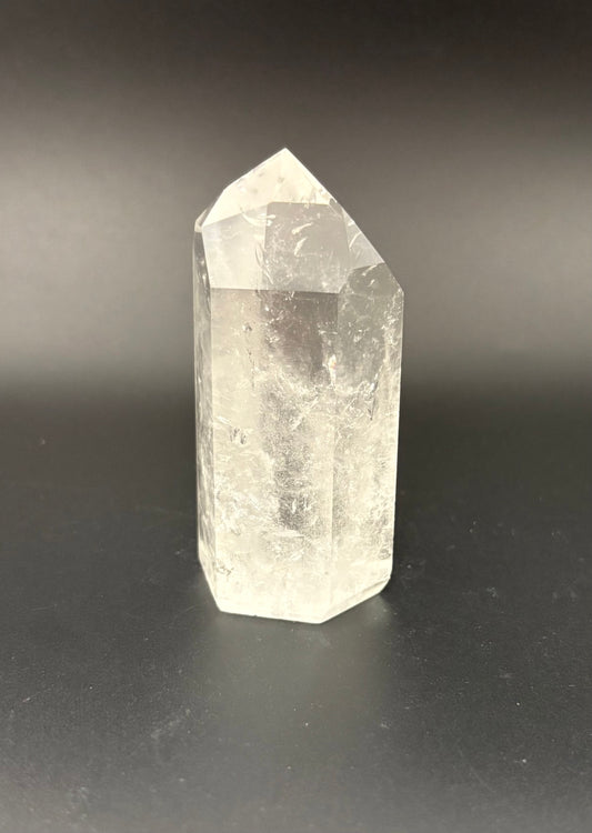A natural clear quartz crystal point tower standing upright on a dark gray surface against a dark gray background.