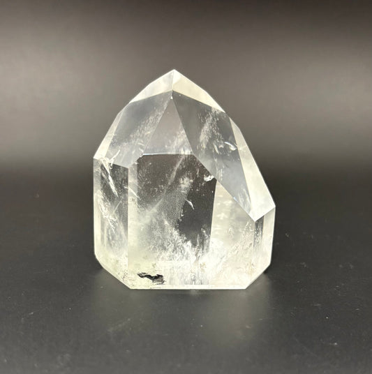 A clear faceted crystal point, quartz, standing on a dark black surface against a dark gray background.