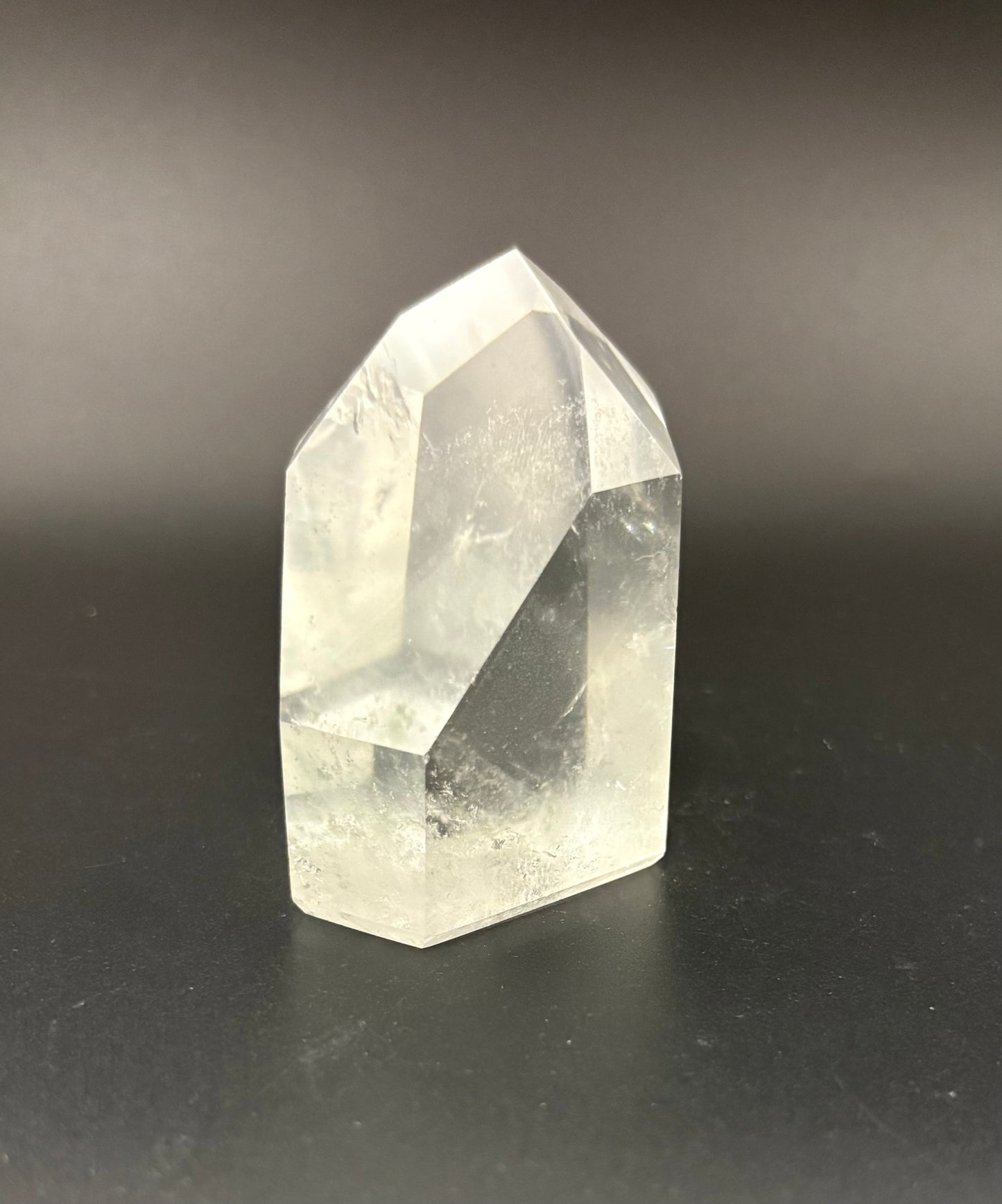 A clear quartz crystal point tower with natural geometric facets, standing on a dark, reflective surface against a dark gray background.