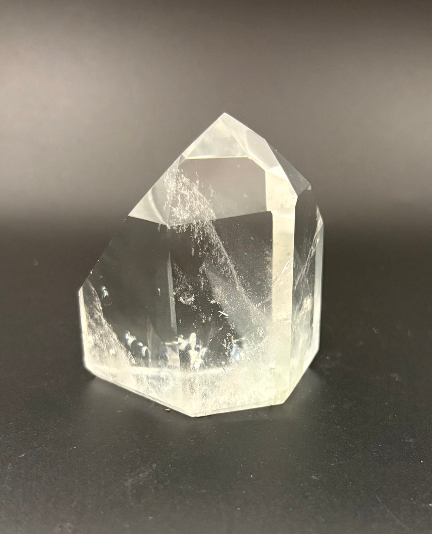 A clear natural quartz crystal point with internal inclusions, standing on a dark gray surface against a dark gray background.