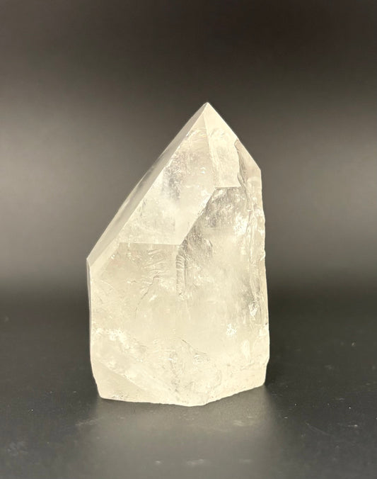 A raw, pointed crystal mineral specimen, likely quartz, showing natural facets and internal inclusions, photographed on a dark gray gradient background.