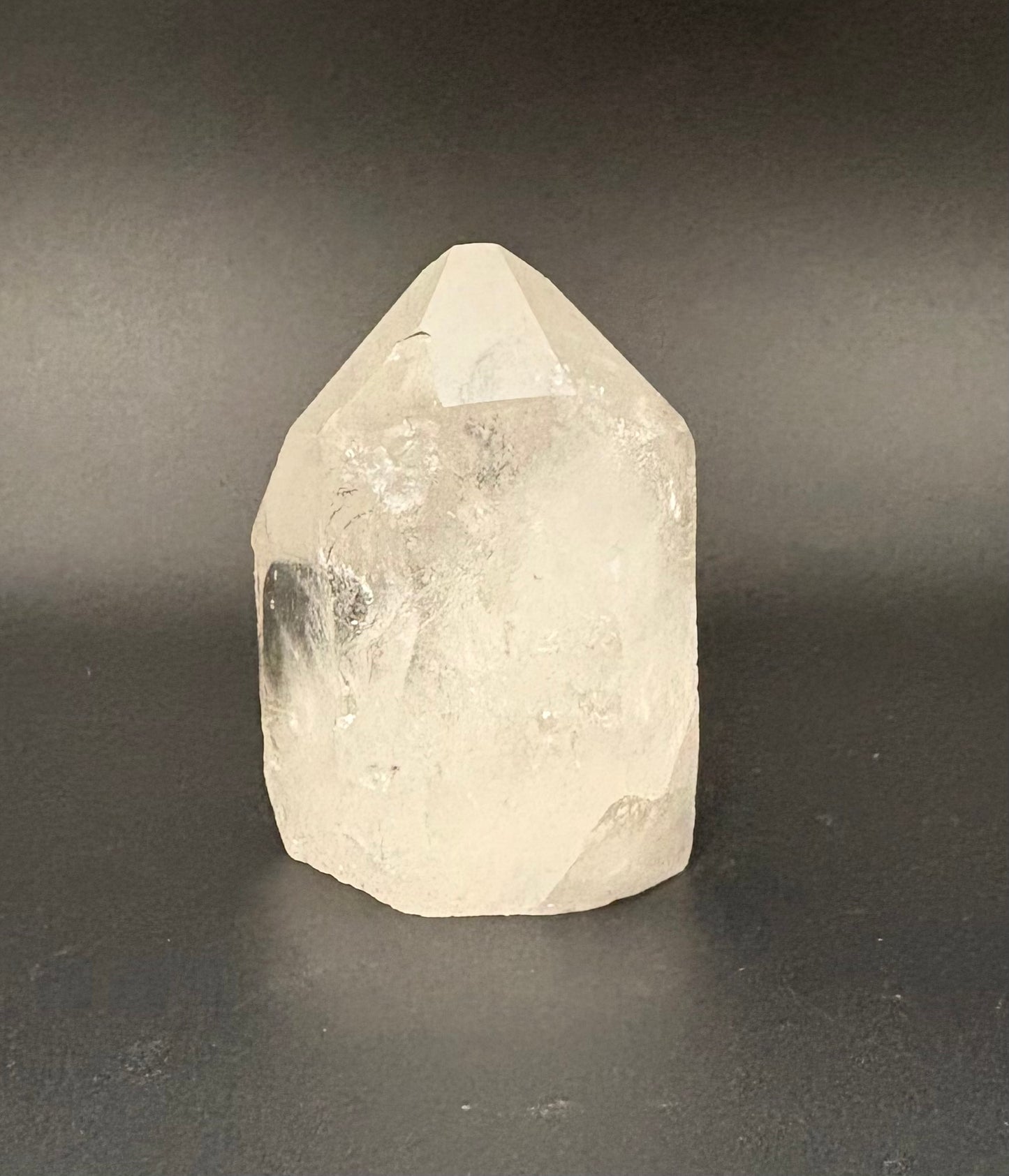 A natural clear quartz point crystal with rough, faceted edges, standing upright on a dark gray surface against a dark gray background.