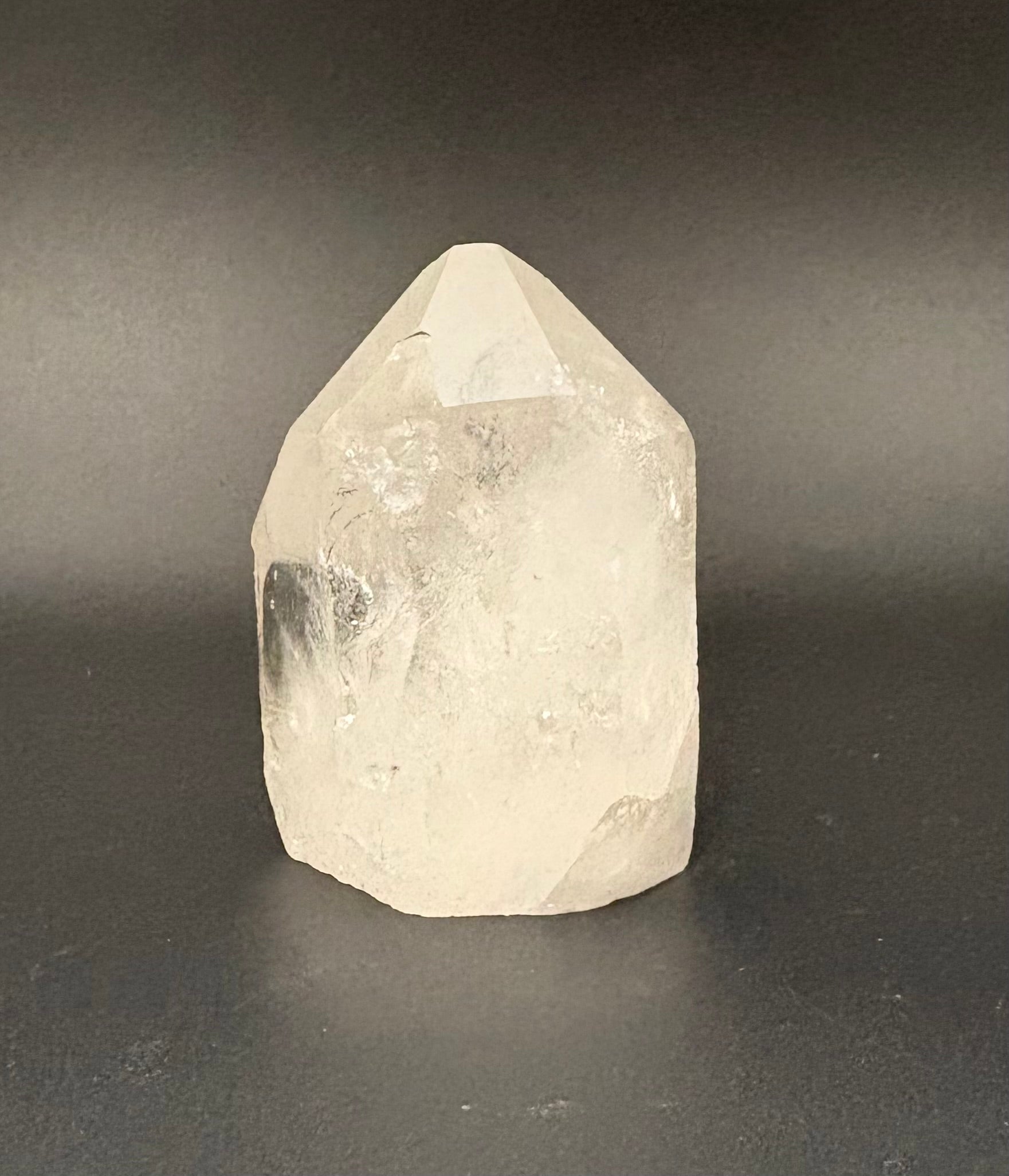 A natural clear quartz point crystal with rough, faceted edges, standing upright on a dark gray surface against a dark gray background.