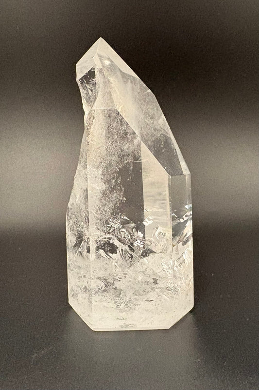 A natural clear quartz crystal tower with sharp geometric facets and internal inclusions, photographed against a dark gray gradient background.