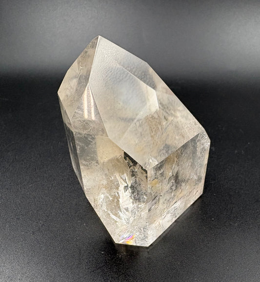 A natural clear quartz crystal tower with sharp geometric facets and internal inclusions, photographed against a dark gray gradient background.