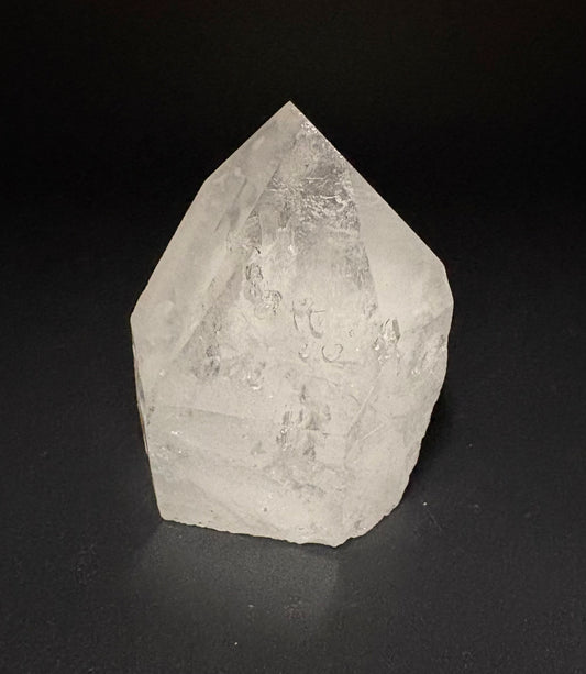 A natural clear quartz crystal point with rough, textured facets, photographed on a dark black background.