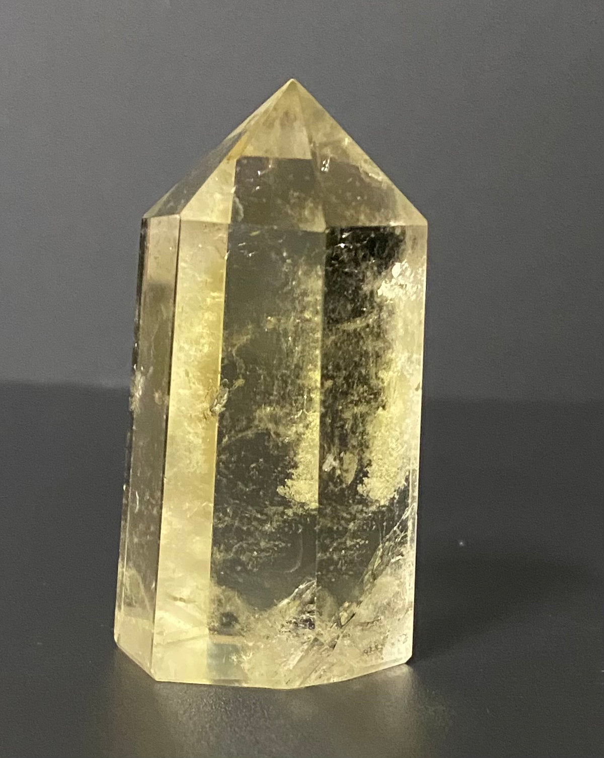 A natural yellow citrine crystal point tower with internal inclusions, standing on a dark gray surface against a dark gray background.