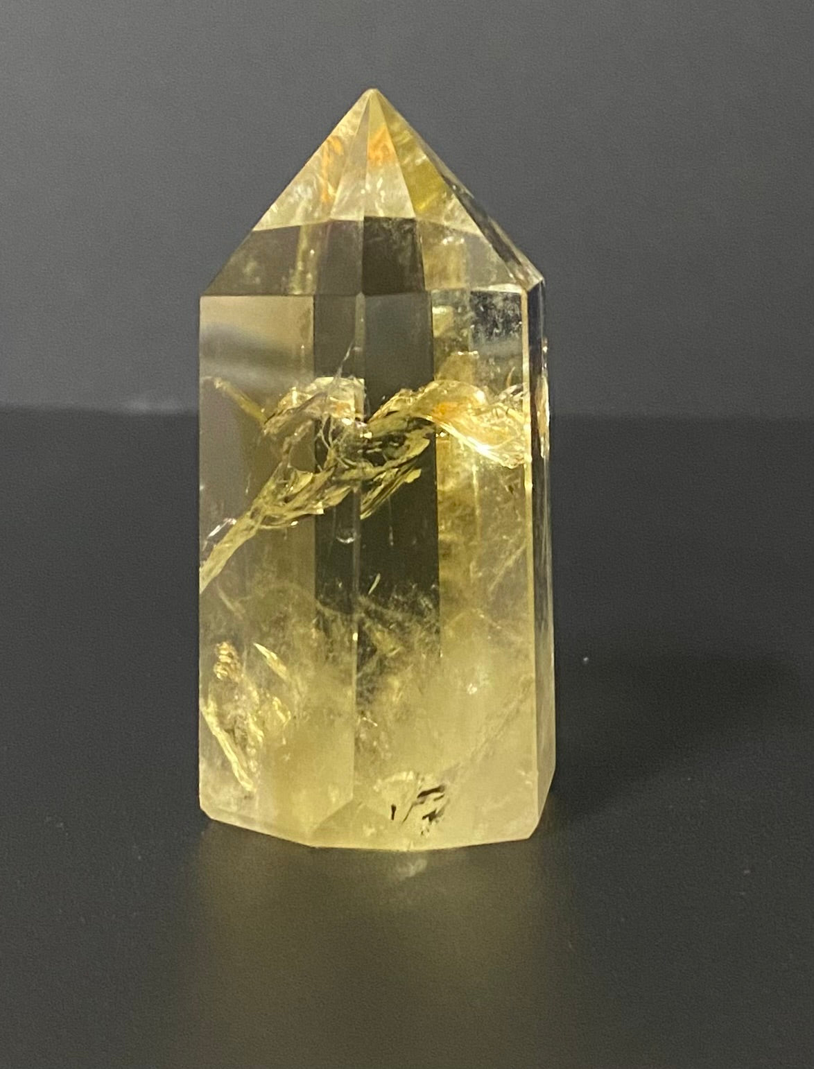 A yellow citrine crystal point tower with natural inclusions, standing on a dark gray surface against a dark gray background.