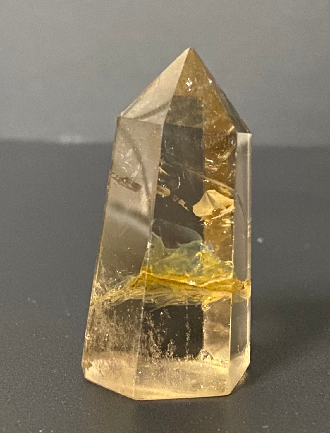 A natural yellow citrine crystal point tower with internal inclusions and a cloudy, semi-translucent appearance, standing on a dark gray surface against a gray background.