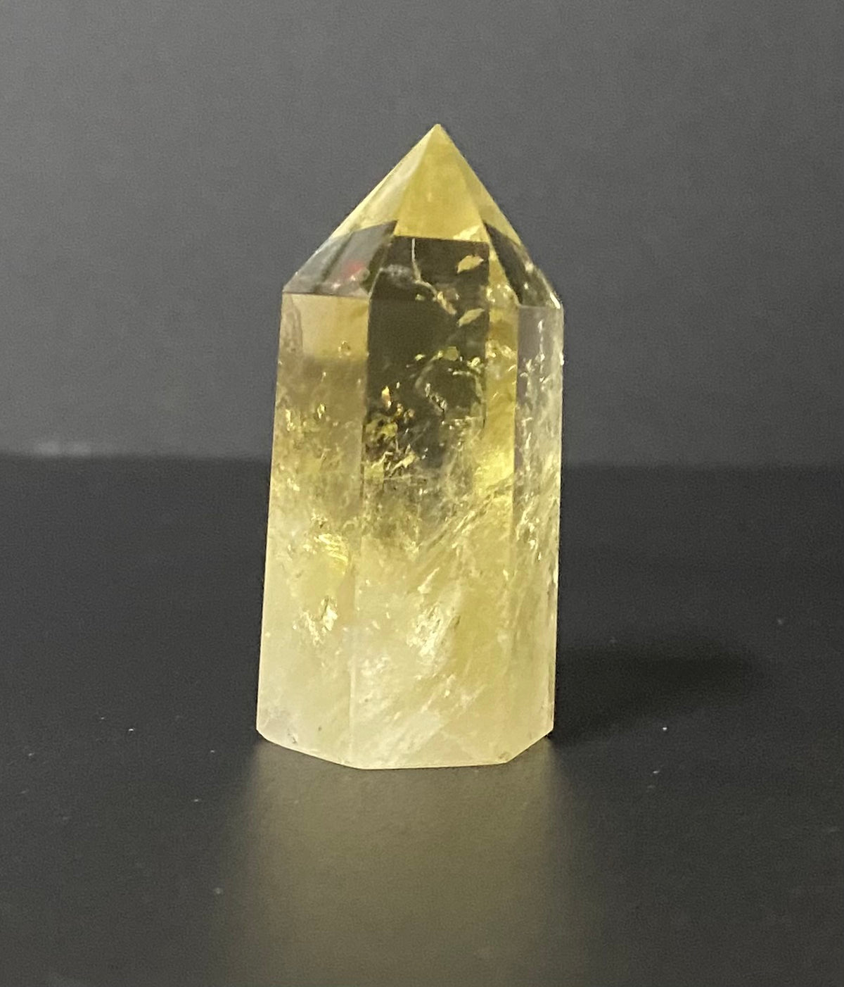 A natural yellow citrine crystal point with internal inclusions, standing on a dark surface against a dark gray background.