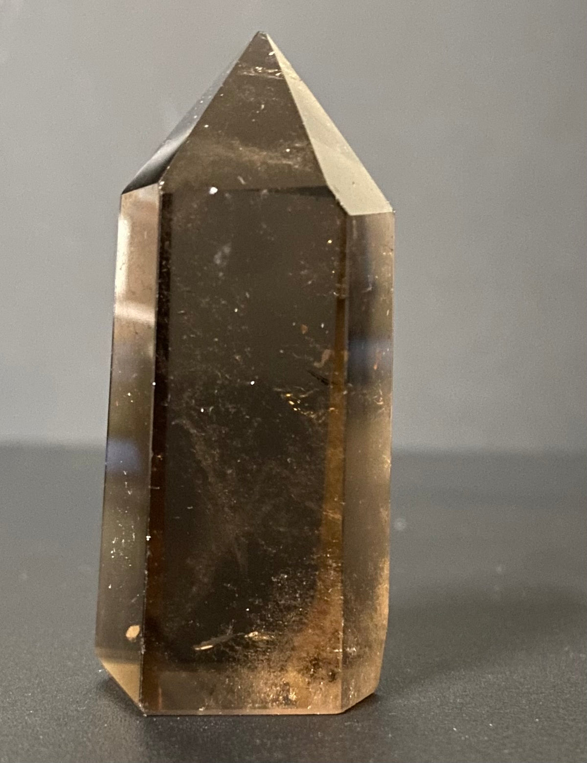 A natural smoky quartz crystal point with a hexagonal shape, showing translucent brown and amber tones with internal inclusions, standing on a dark surface against a neutral gray background.